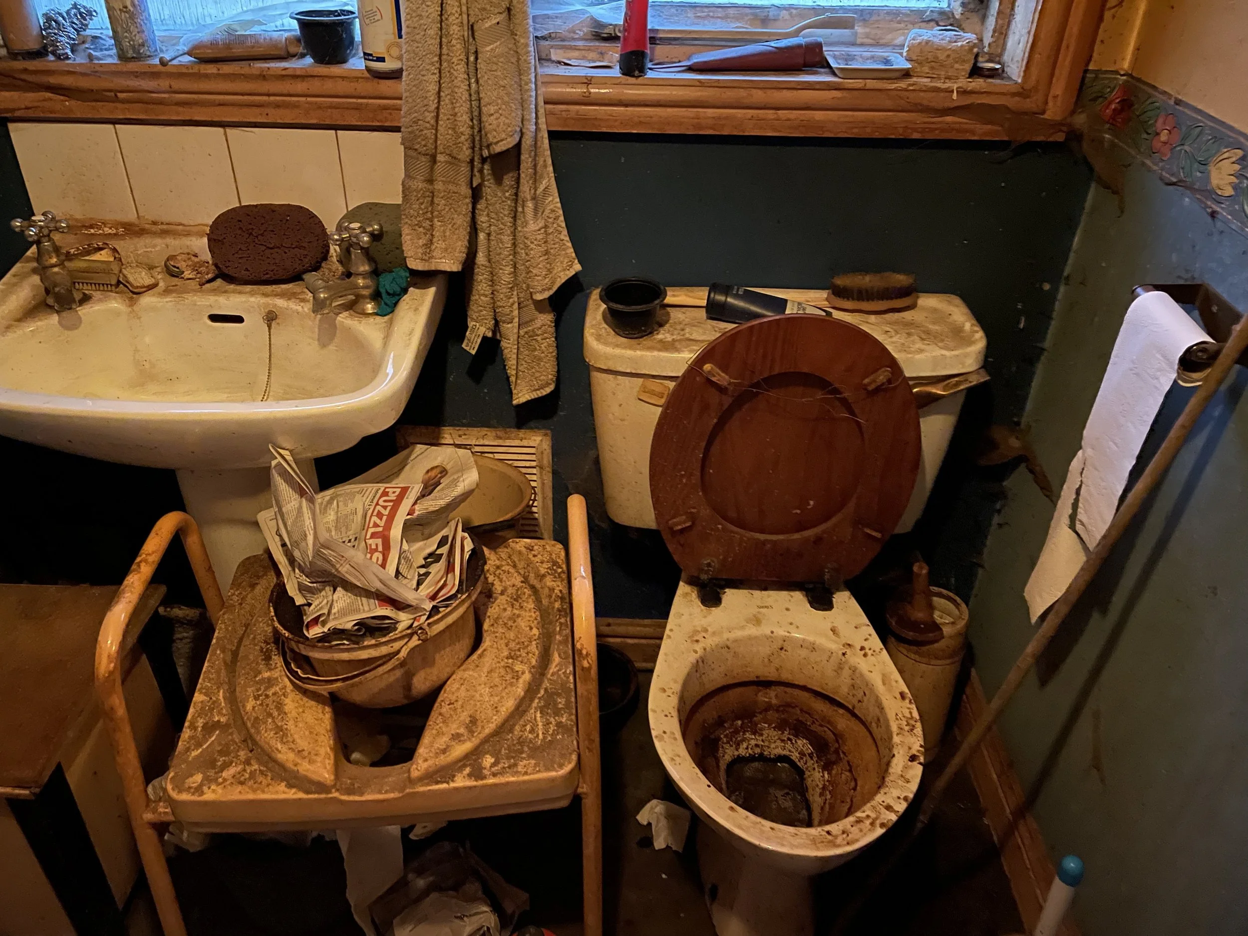 A cluttered bathroom with an old stained sink, a dirty toilet with the lid up, an empty rusty metal basin, a yellowed dresser with disorganized items, and a window with a towel hanging in front of it.