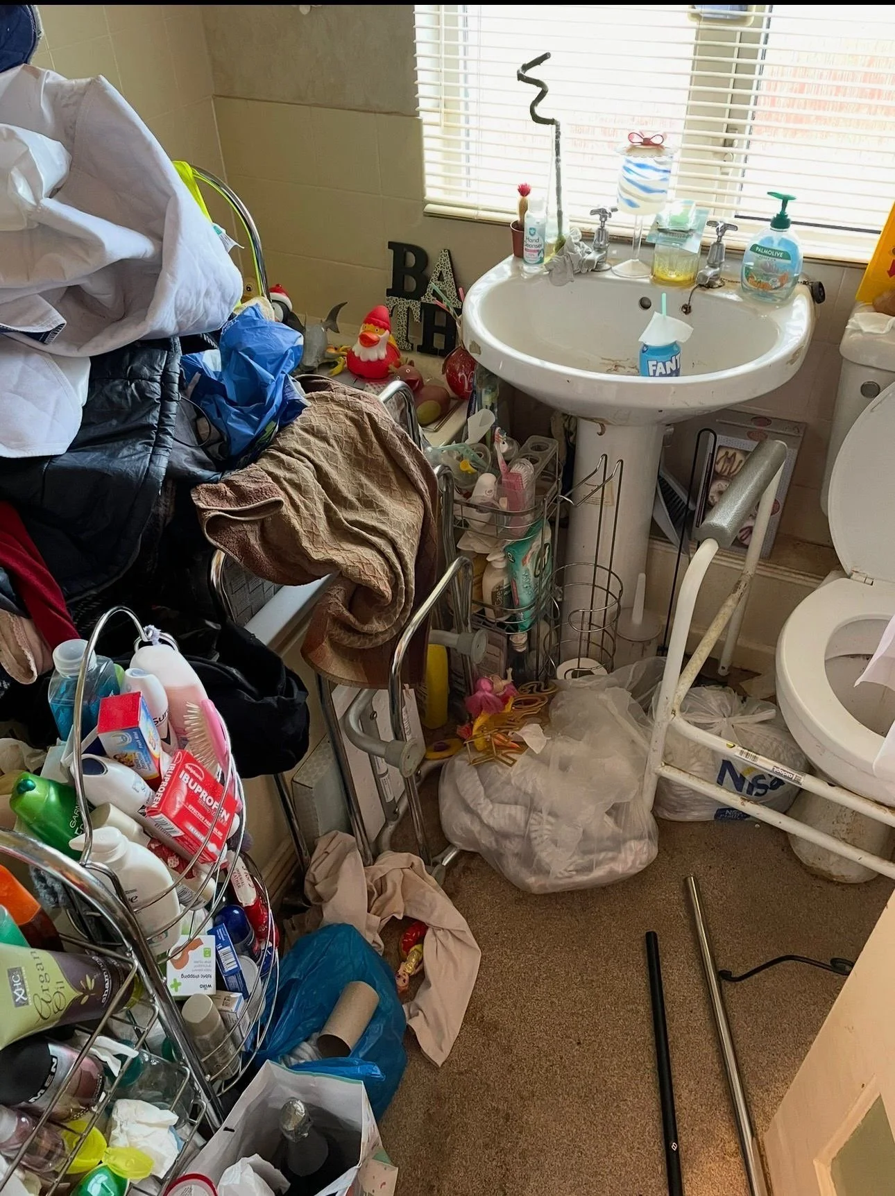 Cluttered bathroom with various toiletries, cleaning supplies, laundry, and miscellaneous items over the sink, shelves, and floor.
