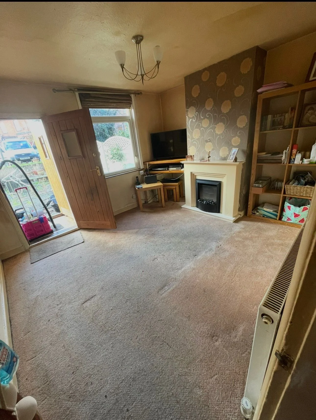Living room with carpet flooring, a wall-mounted television, a fireplace, and a bookshelf. There's an open front door leading outside with a pink plastic bin and broom visible. The room has a ceiling light fixture, and a window lets in natural light.