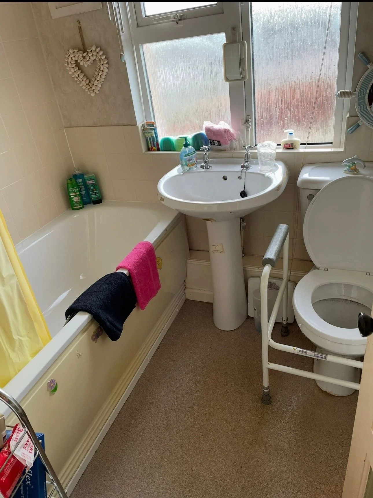A small bathroom with a bathtub, a sink, and a toilet. The window above the sink has a pink cloth and a sponge on the windowsill. Towels hang over the bathtub, and bathroom supplies are on the window ledge and bathtub edge.