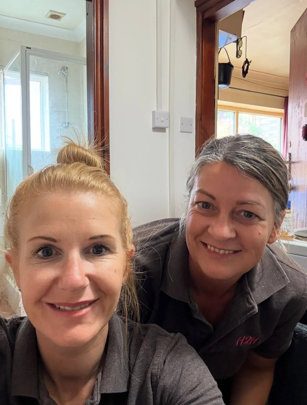 Two women smiling in a room, with a bathroom and window visible in the background.