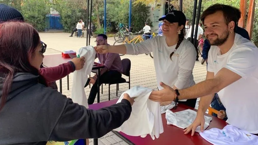 Personas intercambiando un objeto de tela blanca en una mesa al aire libre, con otras personas y bicicletas al fondo.