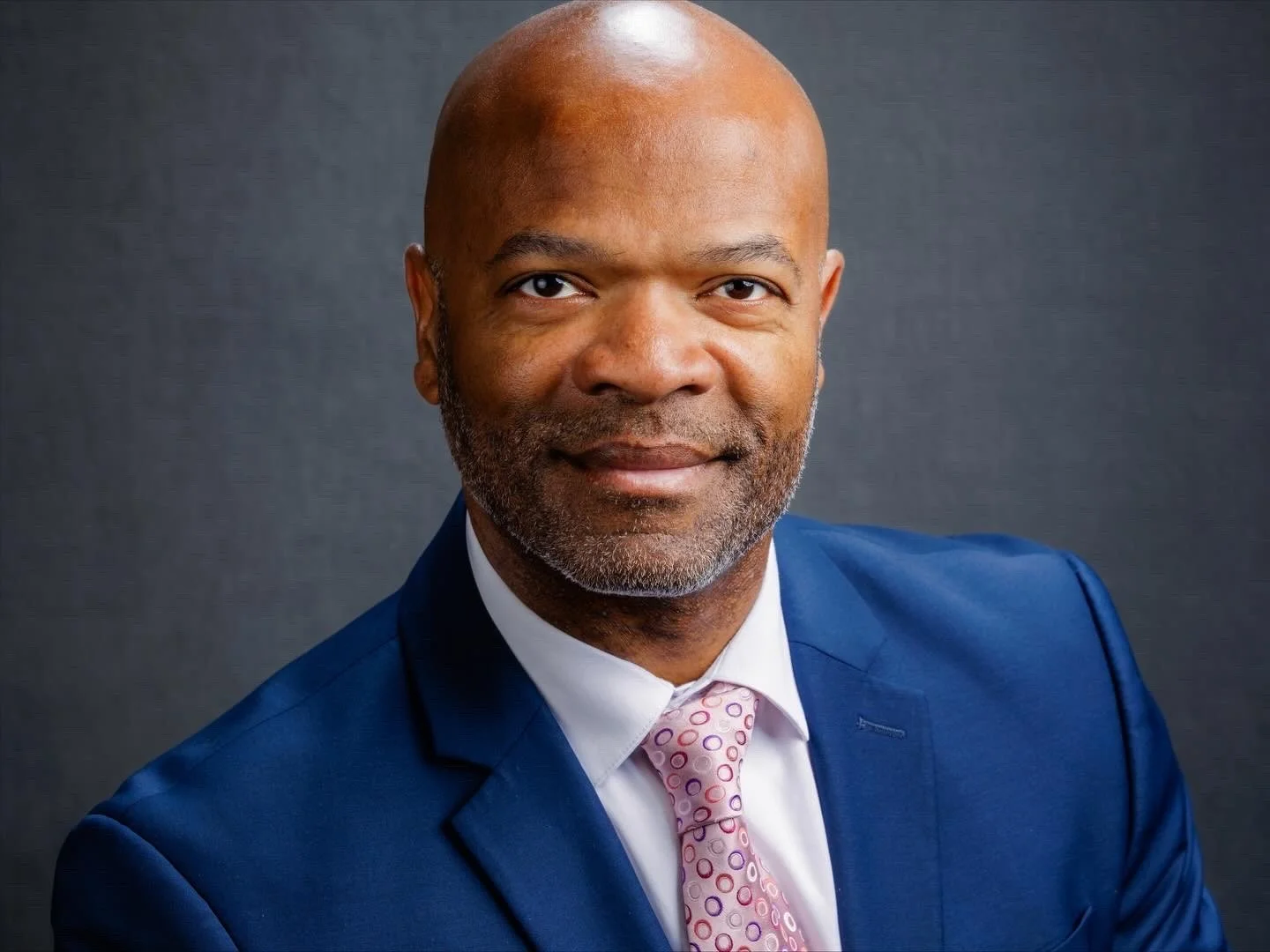 A professional headshot of a man in a blue suit with a white shirt and pink patterned tie, smiling slightly against a dark background.