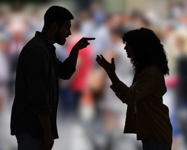 A man and woman engaged in heated argument with fingers pointed at each other, silhouettes against a blurred crowd background.