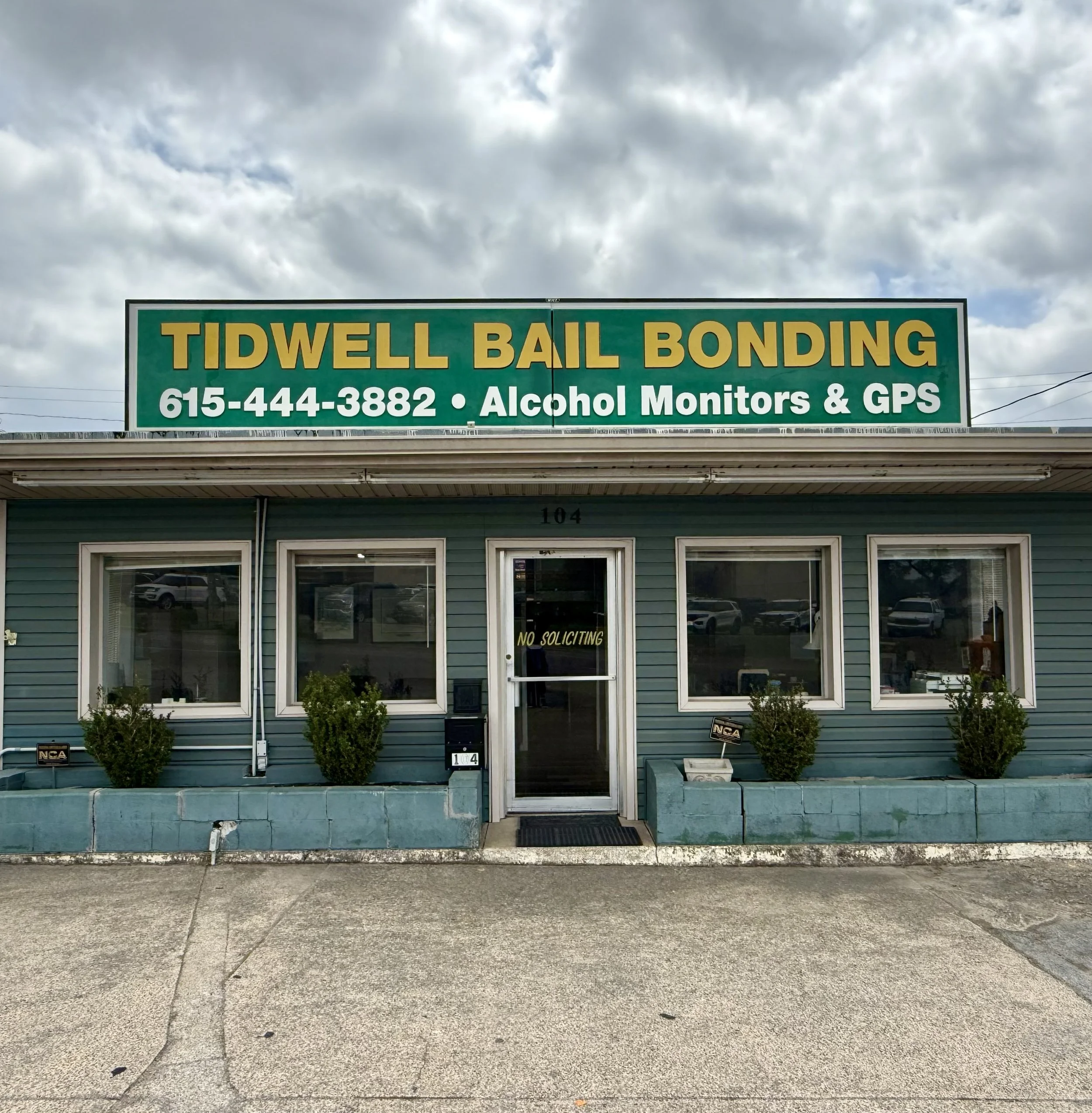 A blue building with three windows and a door in the center, displaying a sign that reads 'No Soliciting.' A green sign on top of the building advertises 'Tidwell Bail Bonding' with a phone number and services like alcohol monitors and GPS. The parking lot in front is cracked, and there are small shrubbery plants in front of the building.