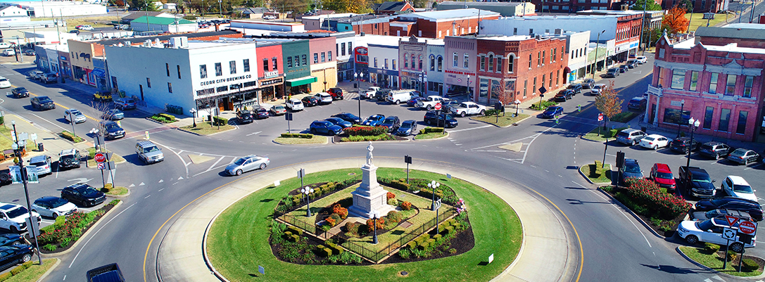 A small town square with a roundabout and a fountain in the center, surrounded by colorful historic buildings and parked cars.