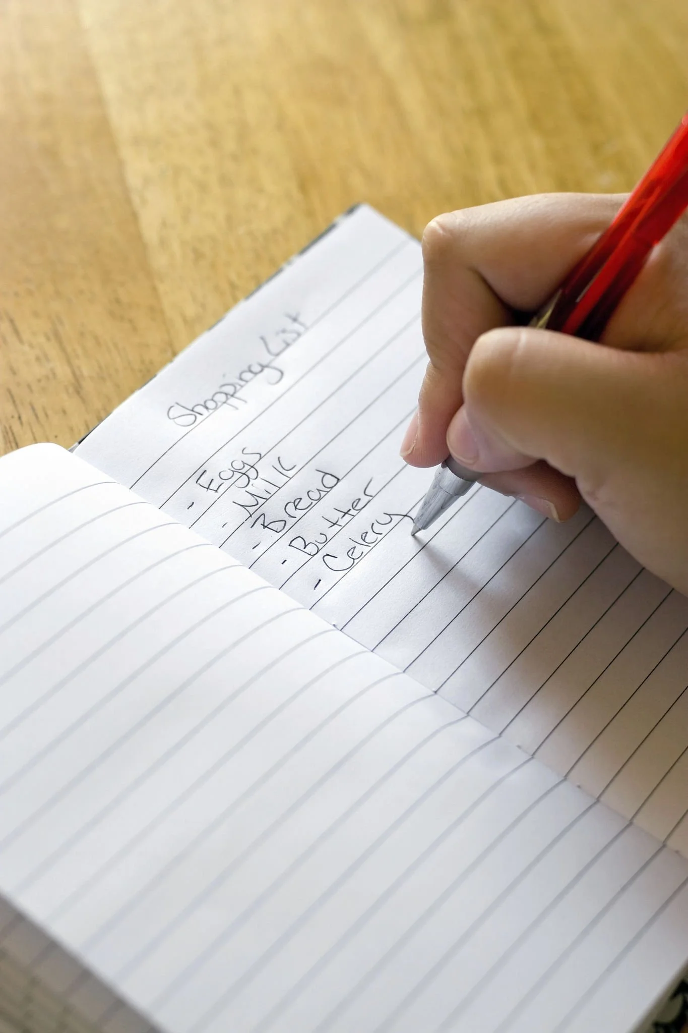 Close-up of a hand writing a grocery list in a lined notebook with a red pen, notes include eggs, milk, bread, butter, and celery.