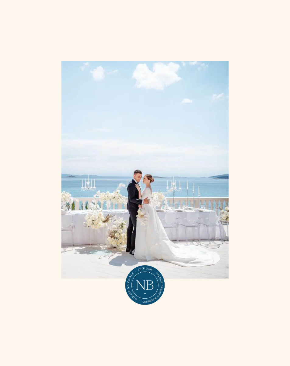 Un couple en robe de mariage célèbre leur mariage en bord de mer avec un décor floral blanc et une vue sur la mer et le ciel bleu.