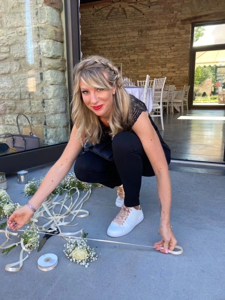 Une femme avec des cheveux blonds et une robe noire, portant un maquillage rouge à lèvres, en train de faire une décoration florale pour un événement dans une salle élégante avec des chaises blanches et une grande fenêtre.