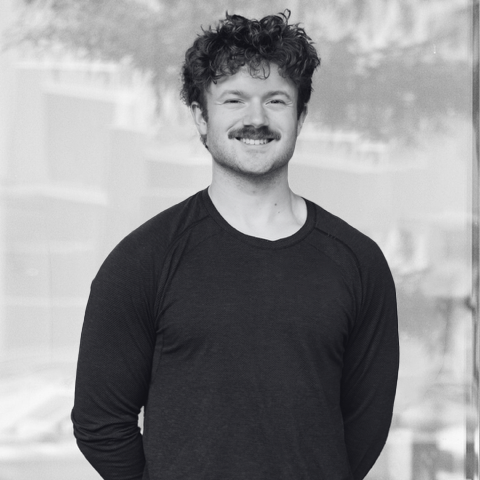 Tanner with curly hair and a mustache smiling, wearing a black long-sleeve shirt, standing outdoors in front of a blurred background.