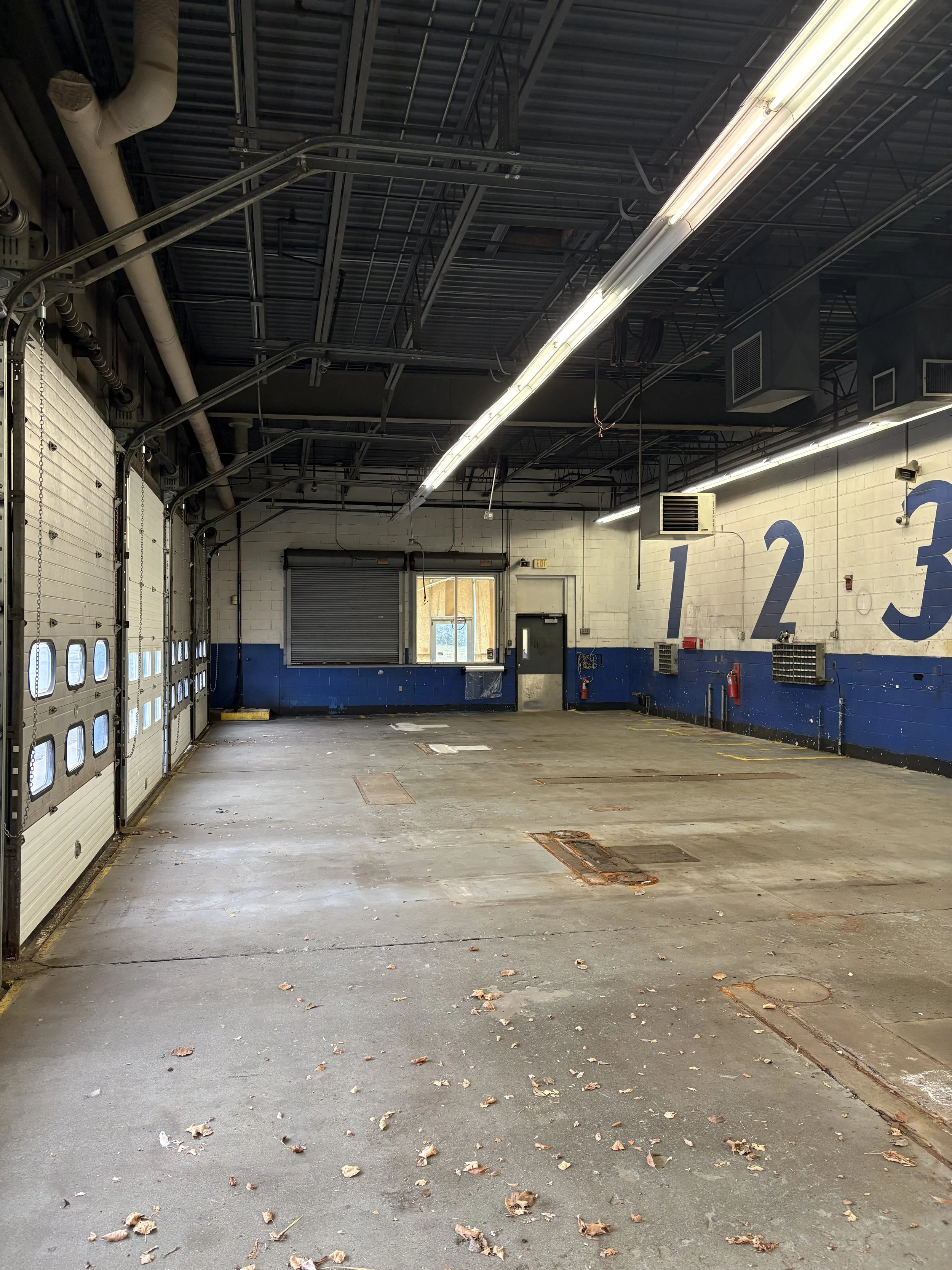 Empty warehouse garage with three large labeled doors numbered 1, 2, and 3, with a small booth window and a door at the back, overhead lights, and debris on the floor.