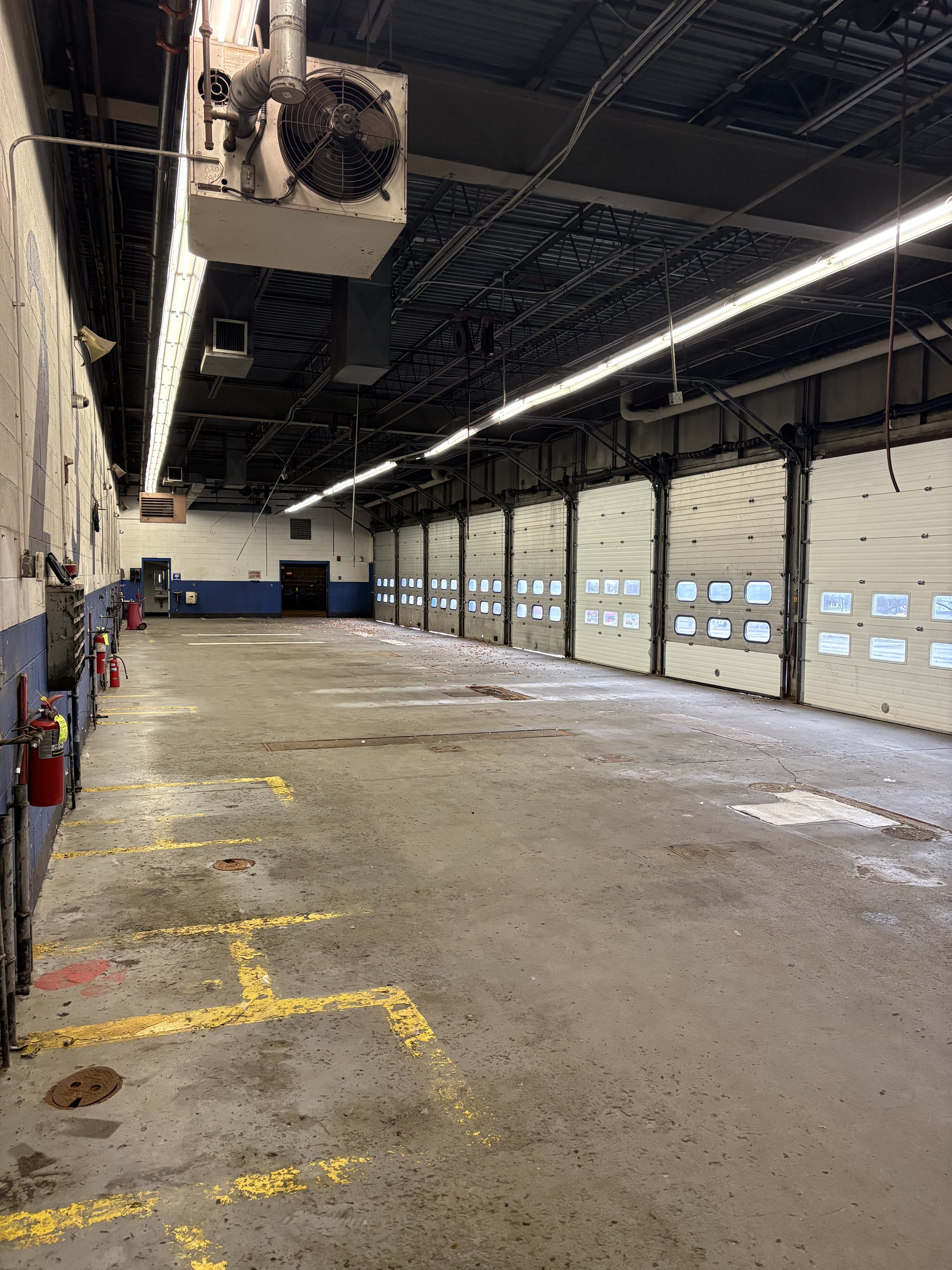 Empty industrial garage or warehouse space with closed garage doors, some yellow parking lines, fire extinguishers on the left wall, and a large ceiling fan.