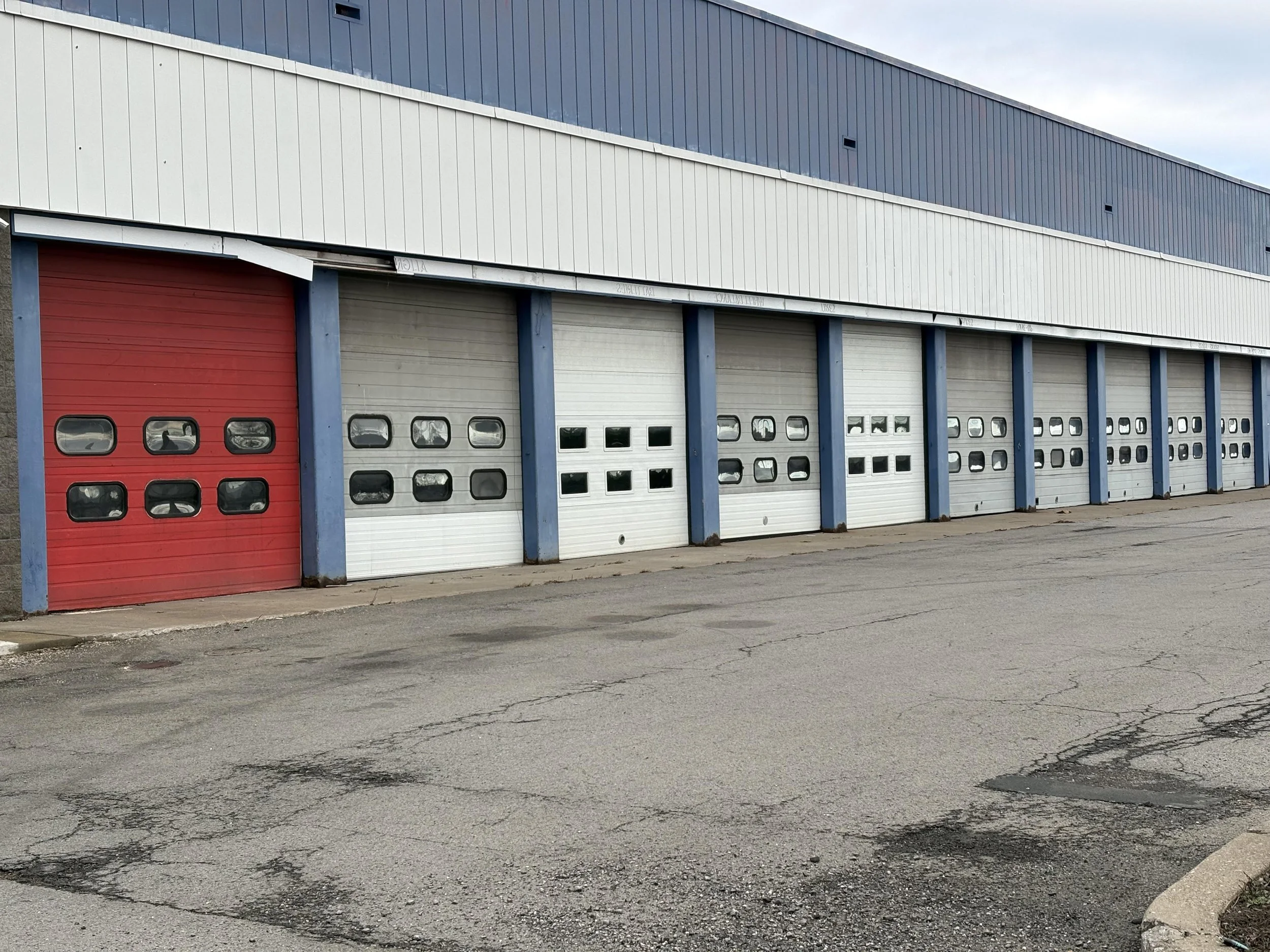 Single-story building with multiple garage doors, one red, the rest white, in an empty parking lot. The building has a blue and white exterior with horizontal metal siding.
