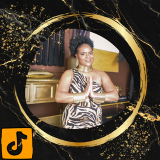 A woman in a zebra print dress and gold jewelry making a prayer pose with her hands together in front of her chest, standing indoors with a yellow pillow and gold furniture.