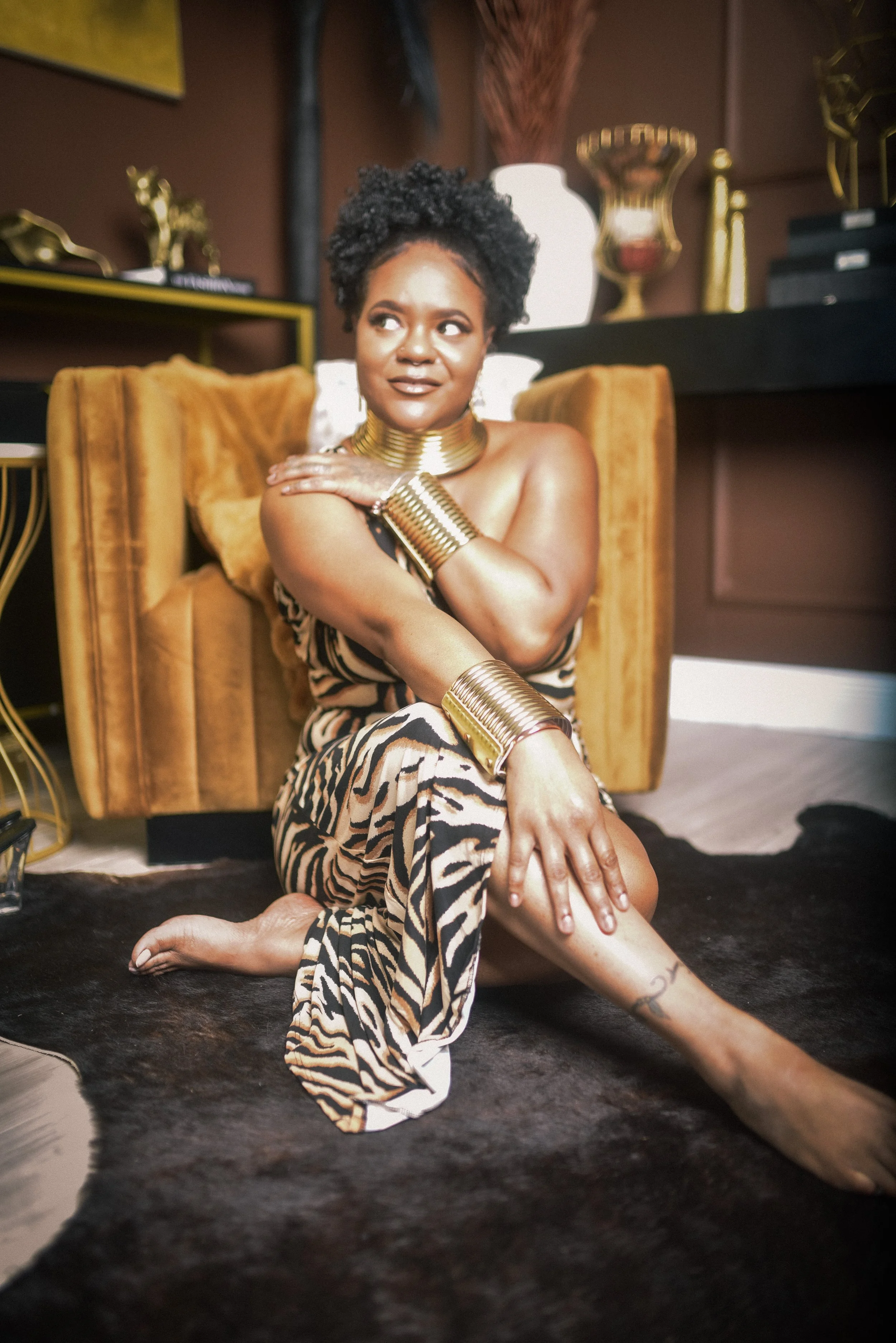 A woman with dark curly hair wearing animal print clothing and large gold jewelry, sitting barefoot on a black shaggy rug in a room with warm lighting and modern decor.