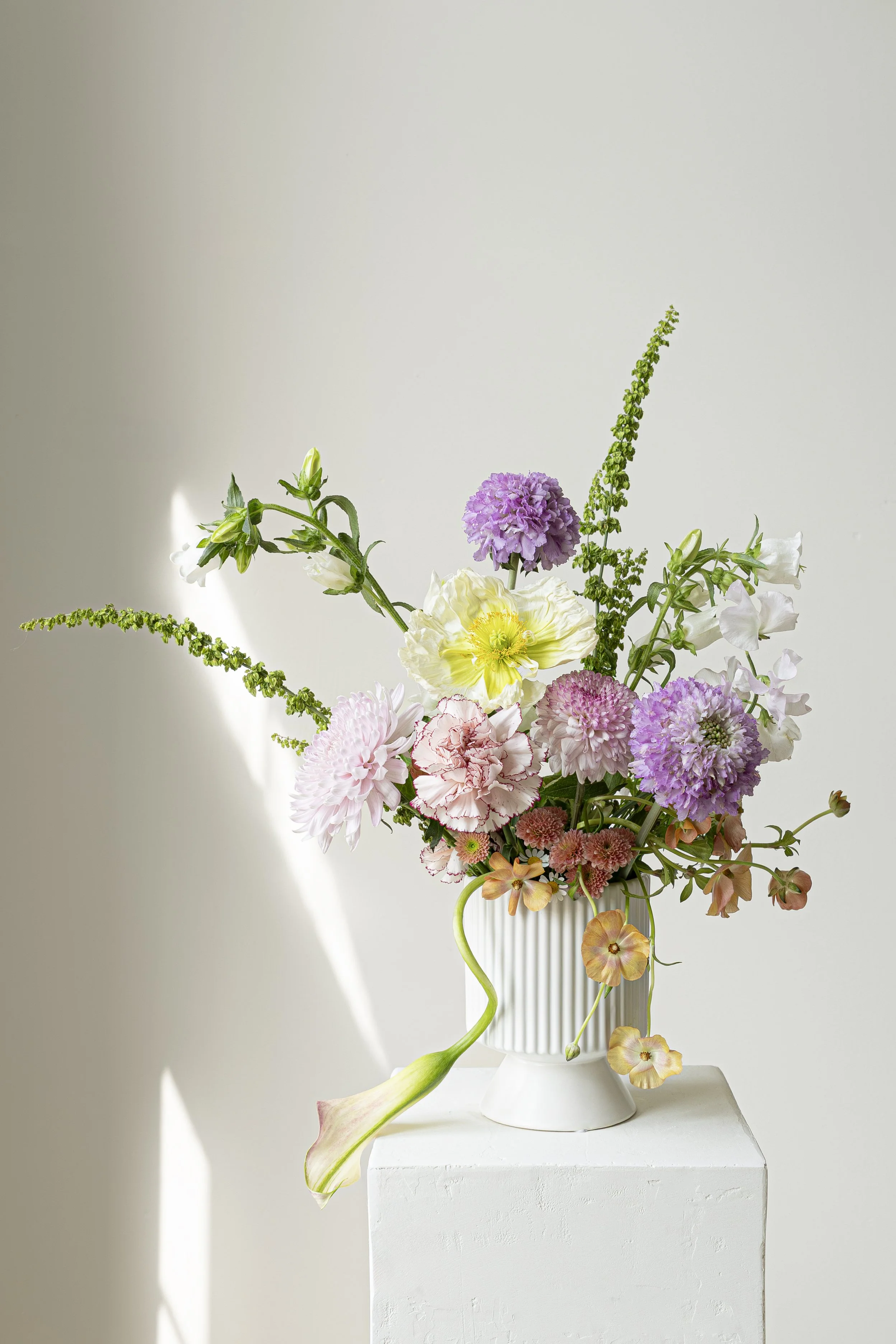 A tall white vase holding a bouquet of various colorful flowers, including pink, purple, white, and orange blossoms, on a white pedestal against a white background with soft natural lighting.