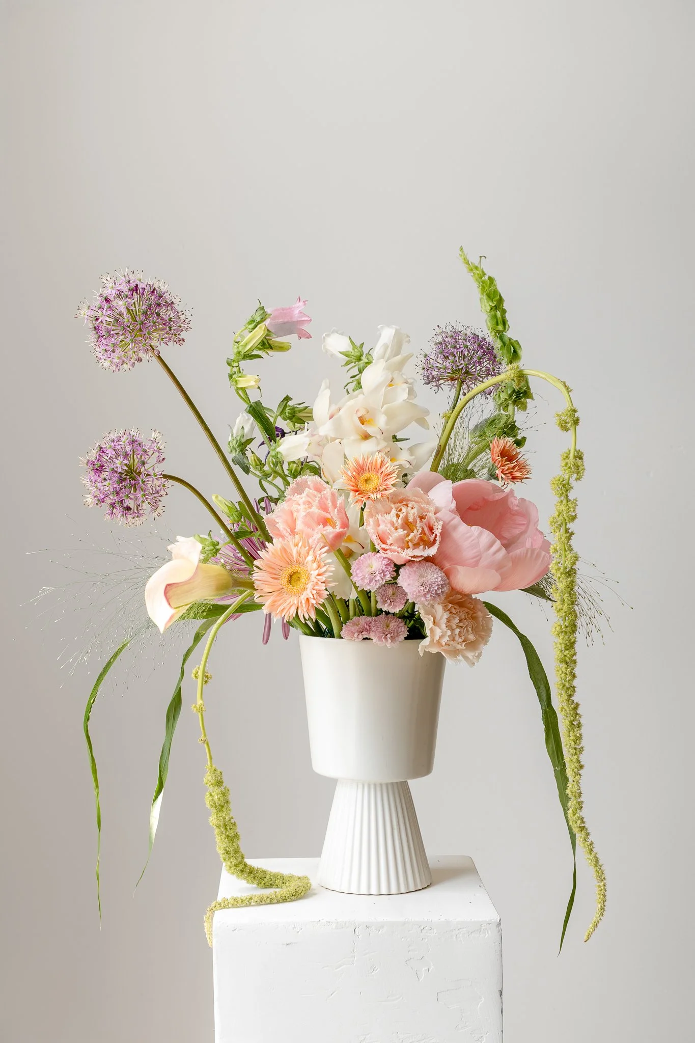 Elegant bouquet of pink, white, and purple flowers in a white vase on a white pedestal against a plain light gray background.