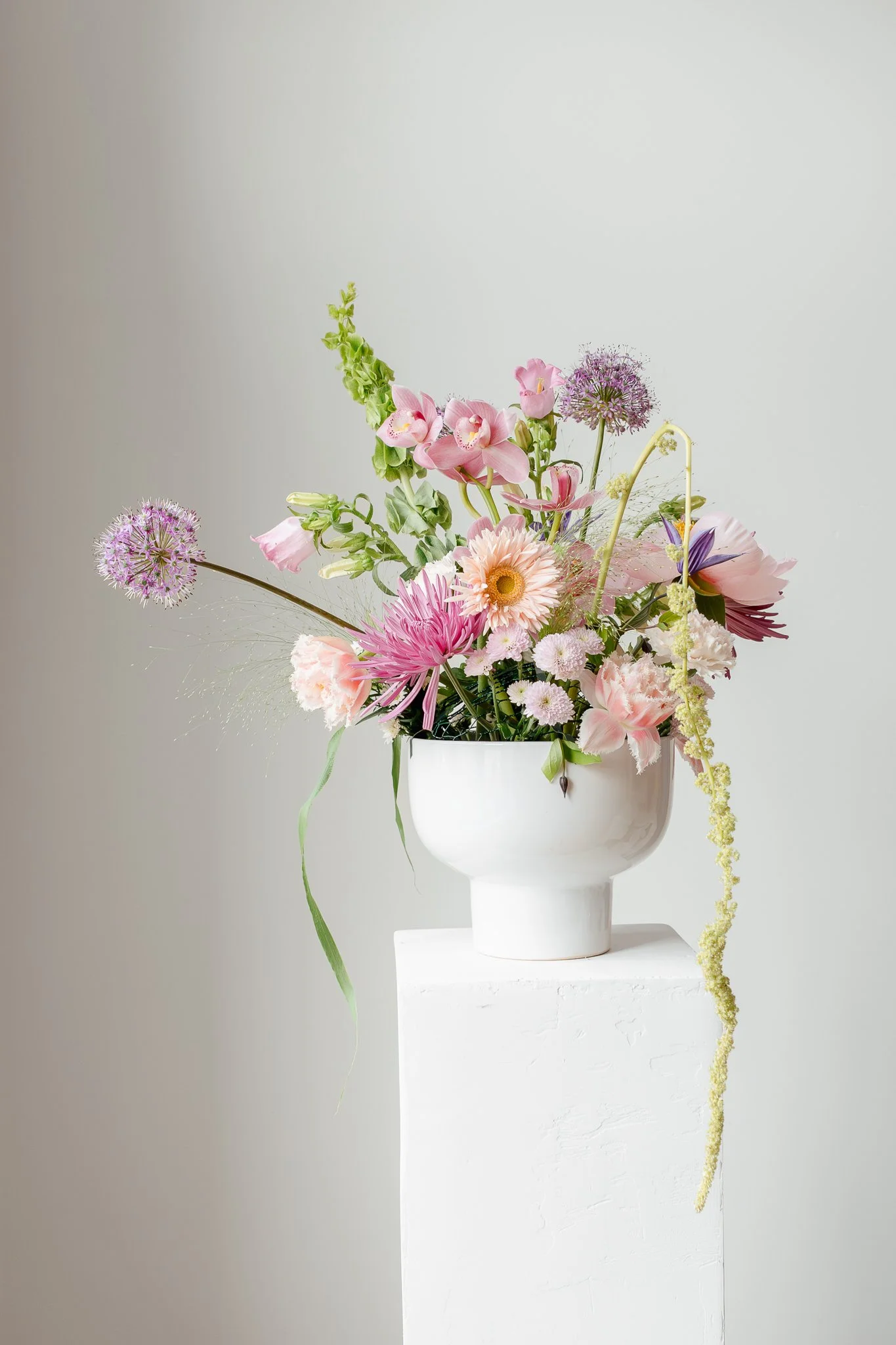 A flower arrangement with pink, purple, and white flowers in a white vase, placed on a white pedestal against a plain light background.
