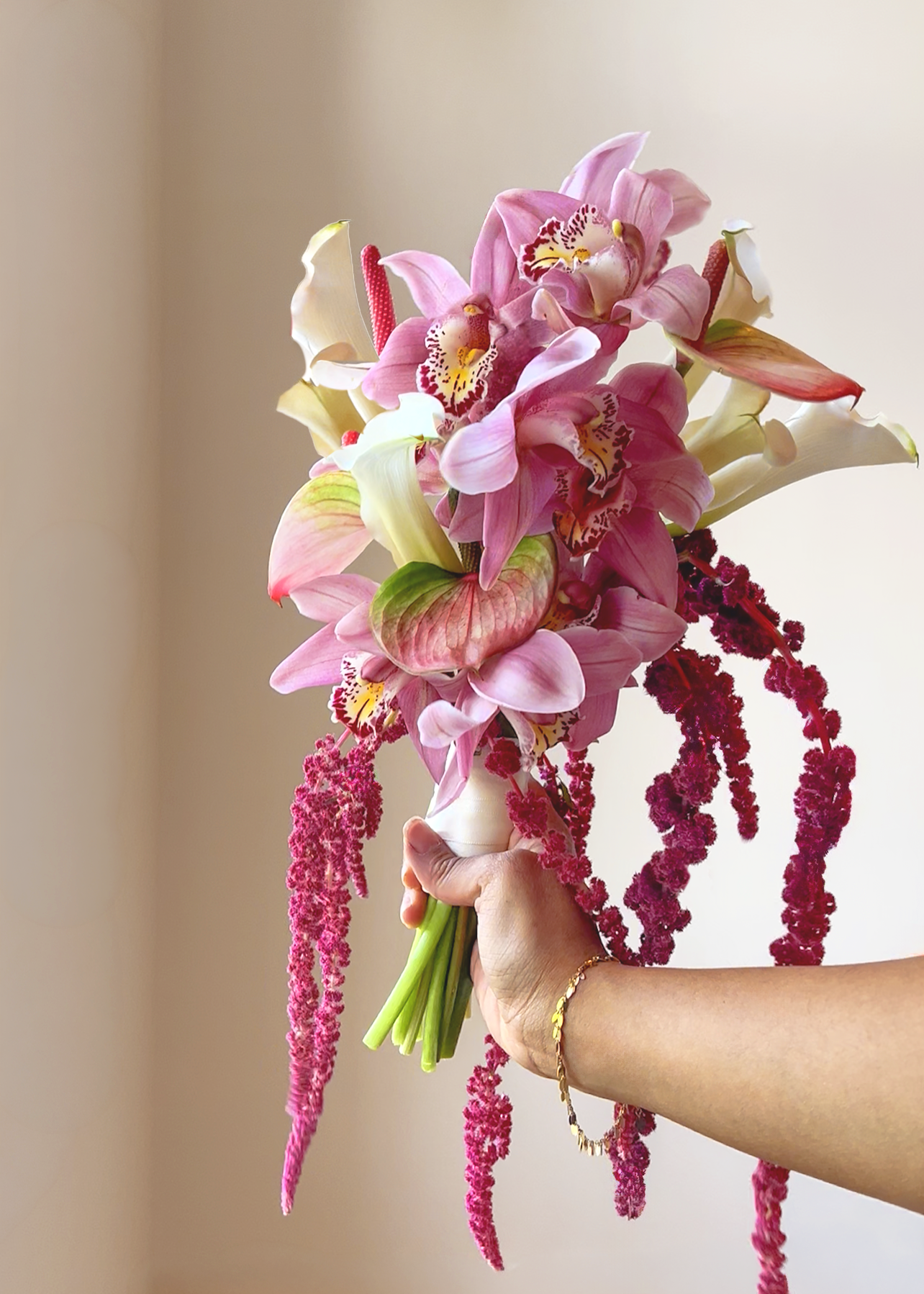 A hand holding a vibrant bouquet of pink and cream orchids, with hanging pink amaranthus, against a neutral background.