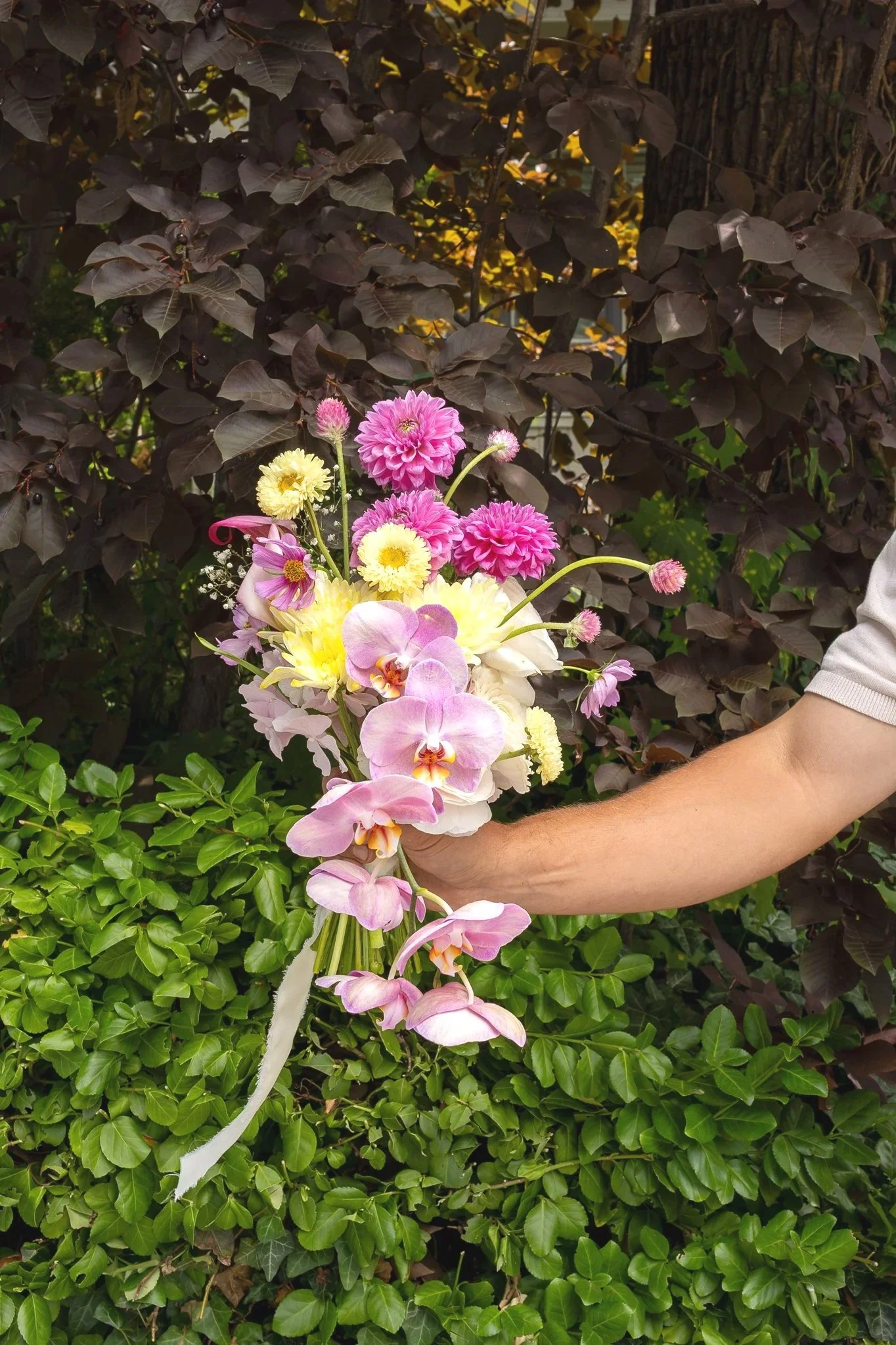 Close-up of a person holding a bouquet of pink, yellow, and white flowers in front of a dark leafy background with plants and trees.