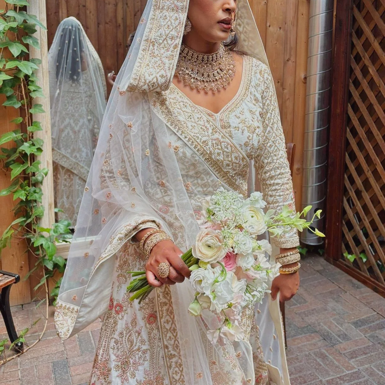 A woman dressed in traditional wedding attire, holding a bouquet of white and pale pink flowers, standing in a wooden outdoor setting.