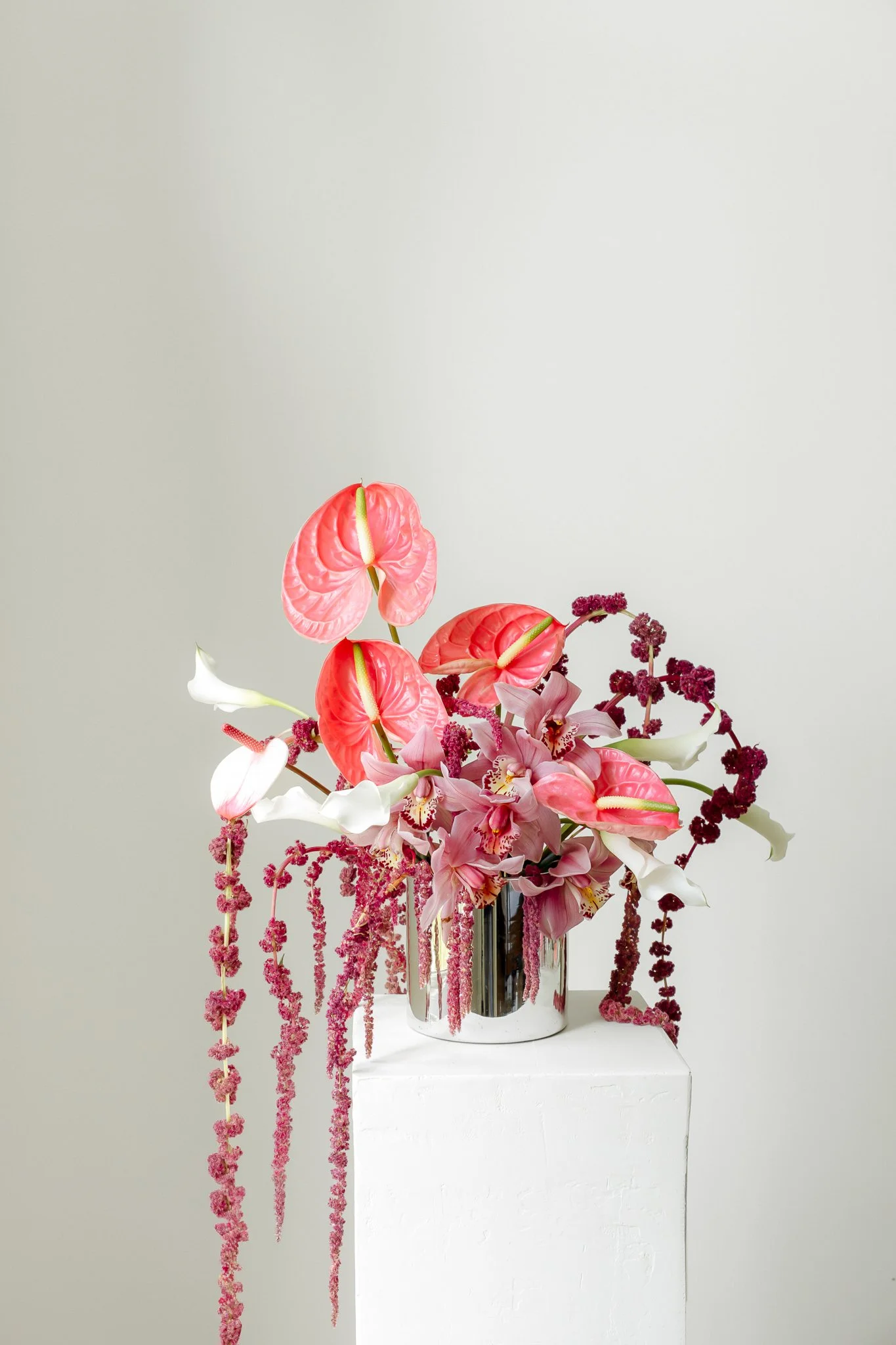 A floral arrangement in a silver vase with pink anthuriums, white calla lilies, orchids, and hanging pink amaranthus, displayed on a white pedestal against a plain light gray background.