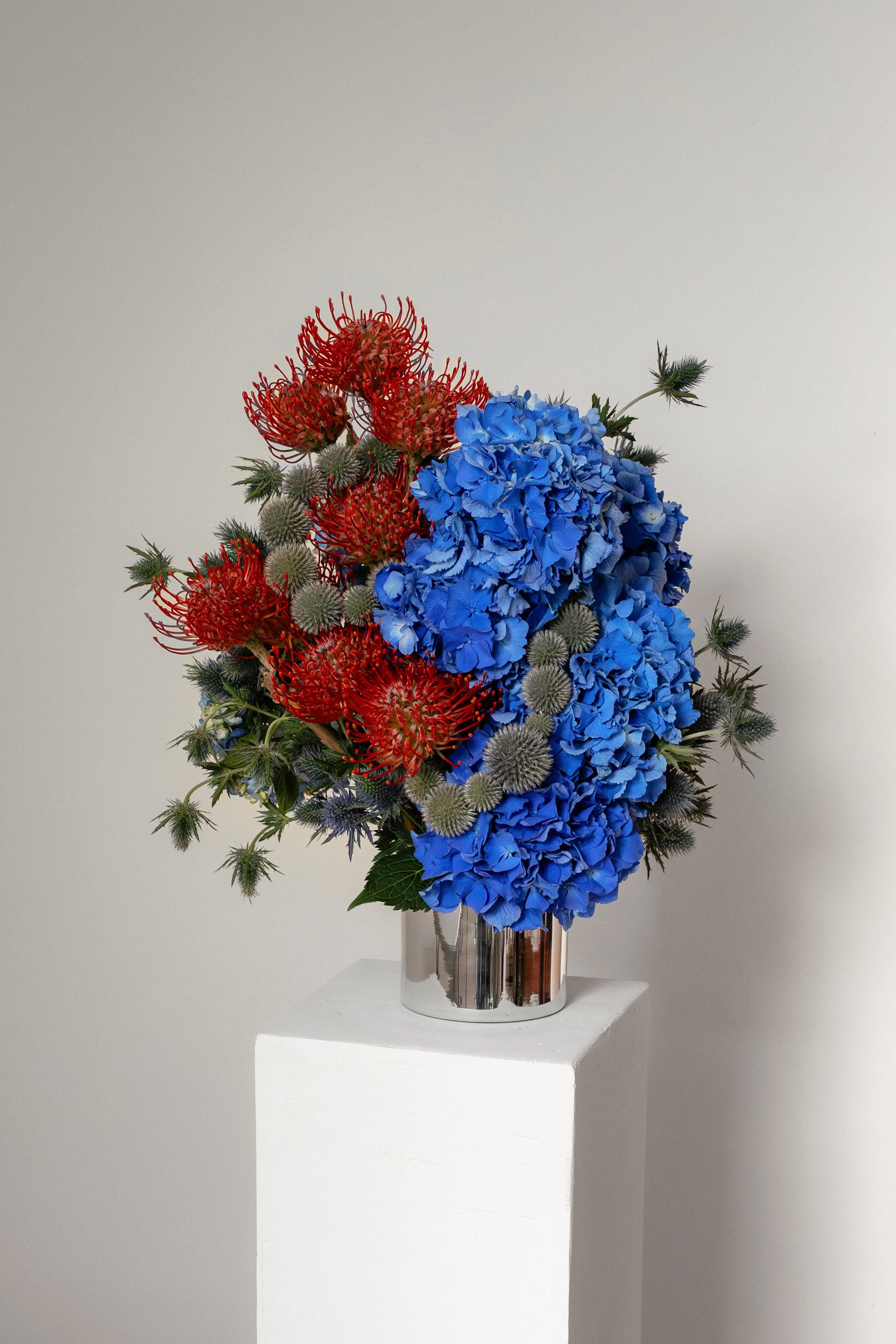 A floral arrangement in a shiny metallic vase on a white pedestal against a gray wall, featuring red, blue, and grayish-green flowers and foliage.