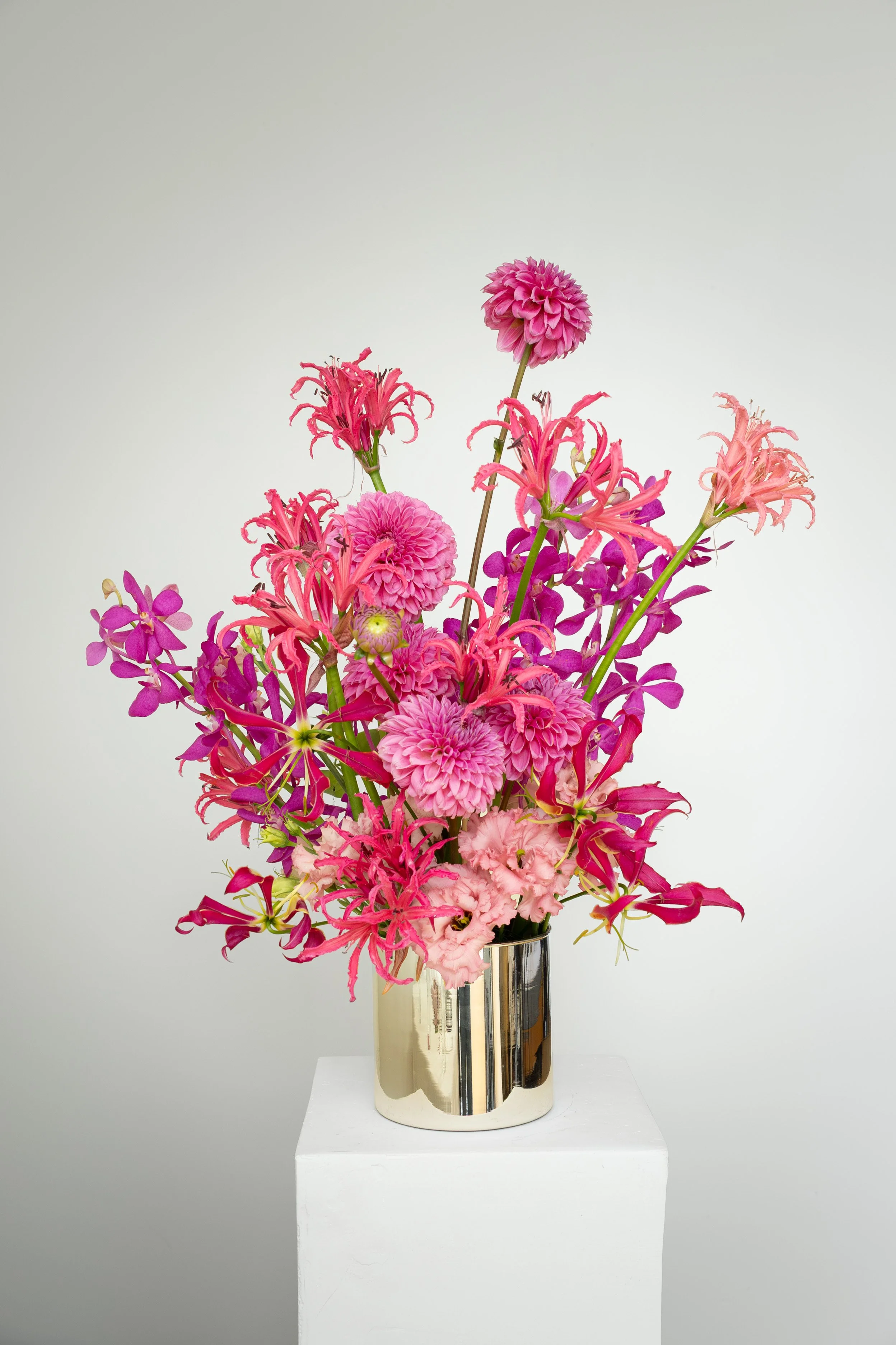 Arrangement of pink and purple flowers in a gold and silver vase on a white pedestal against a plain white background.