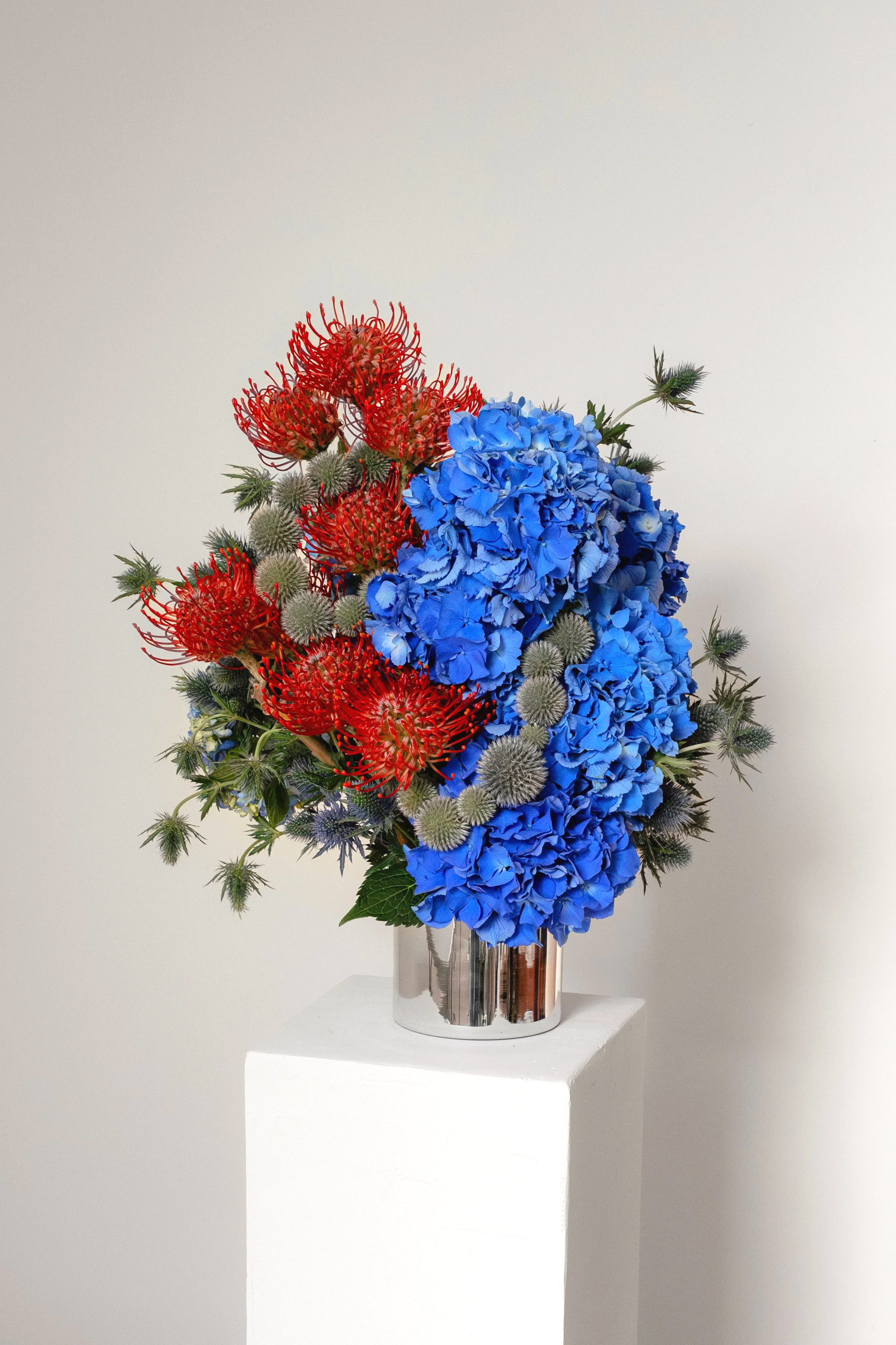 A bouquet of blue hydrangeas and red flowers arranged in a shiny silver vase, displayed on a white pedestal against a plain light background.