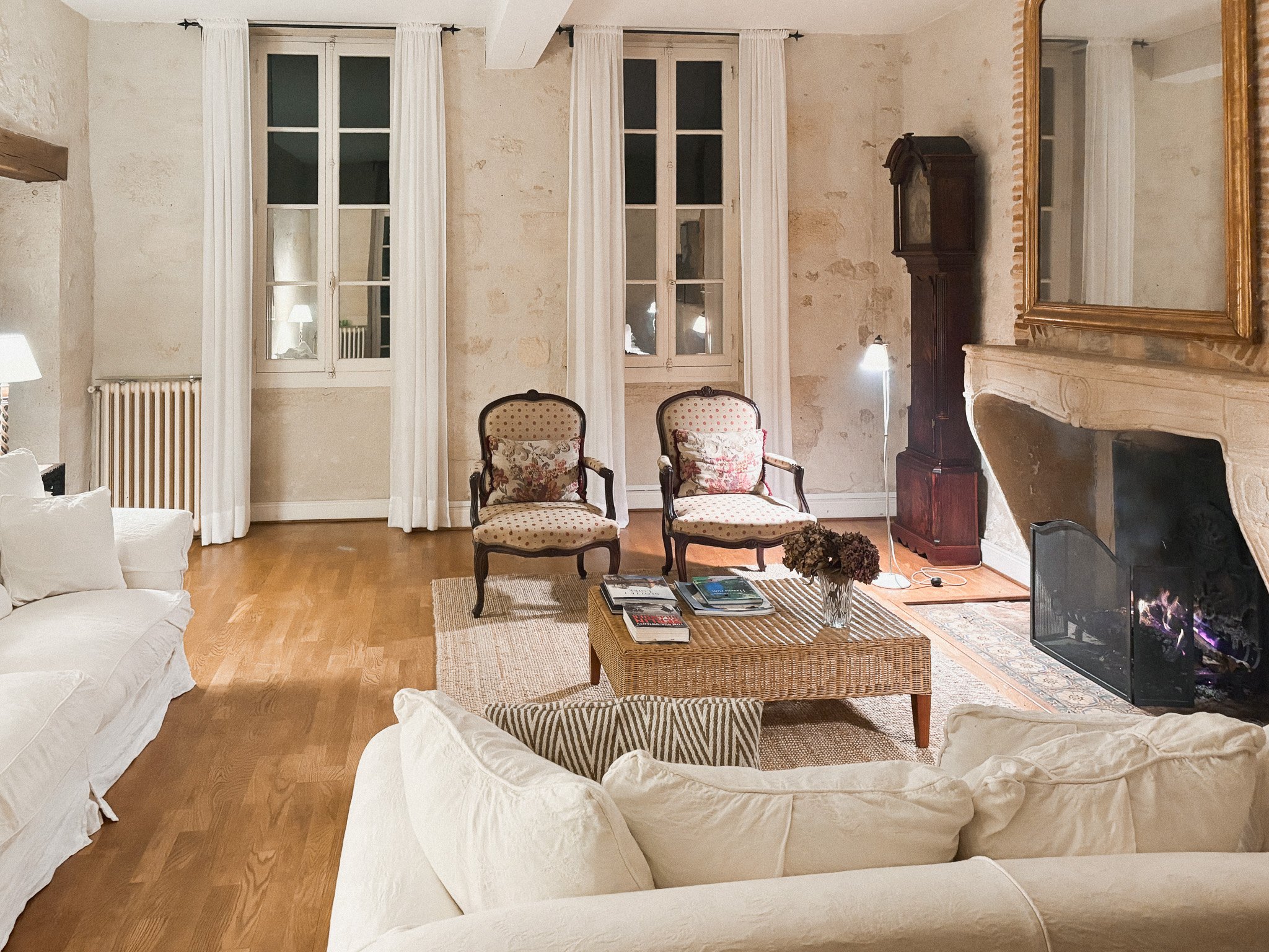 Living room with beige stone walls, large windows with white curtains, a white couch, two vintage armchairs with floral cushions, a wooden coffee table with magazines and flowers, a fireplace with a fire burning, and a tall mirror above the mantel.