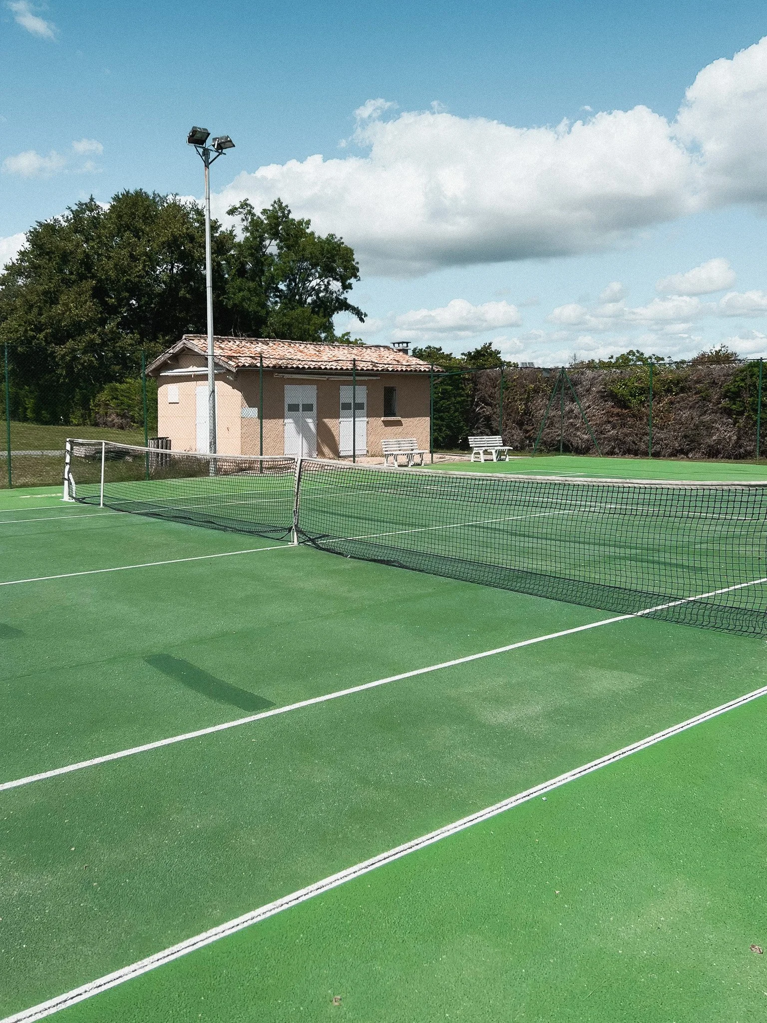 Empty green tennis courts with a net in the center, a small beige building, benches, a tall light pole, trees, and a partly cloudy sky.