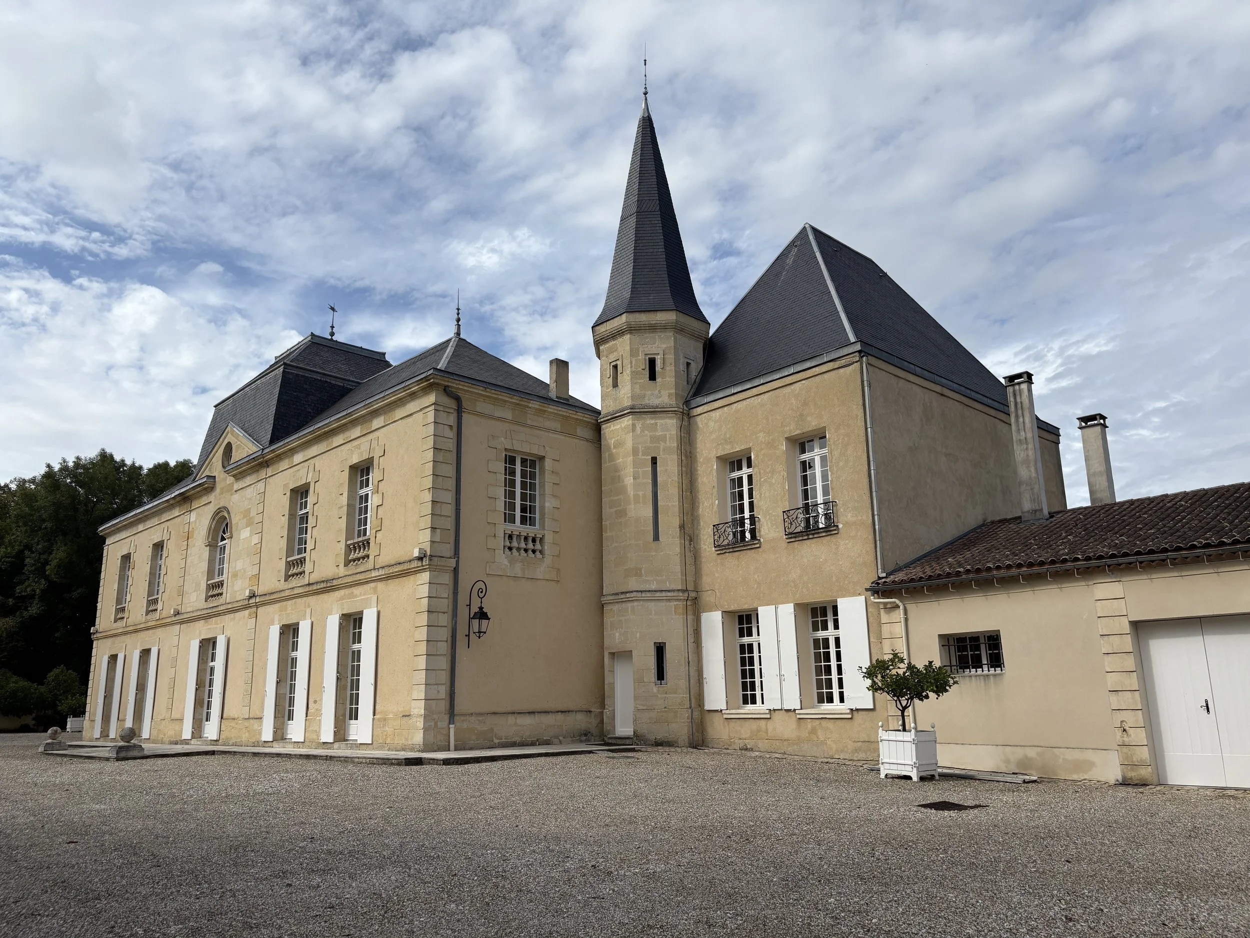 A historic castle with beige stone walls, dark slate roofs, and a tall pointed turret, set against a partly cloudy sky.