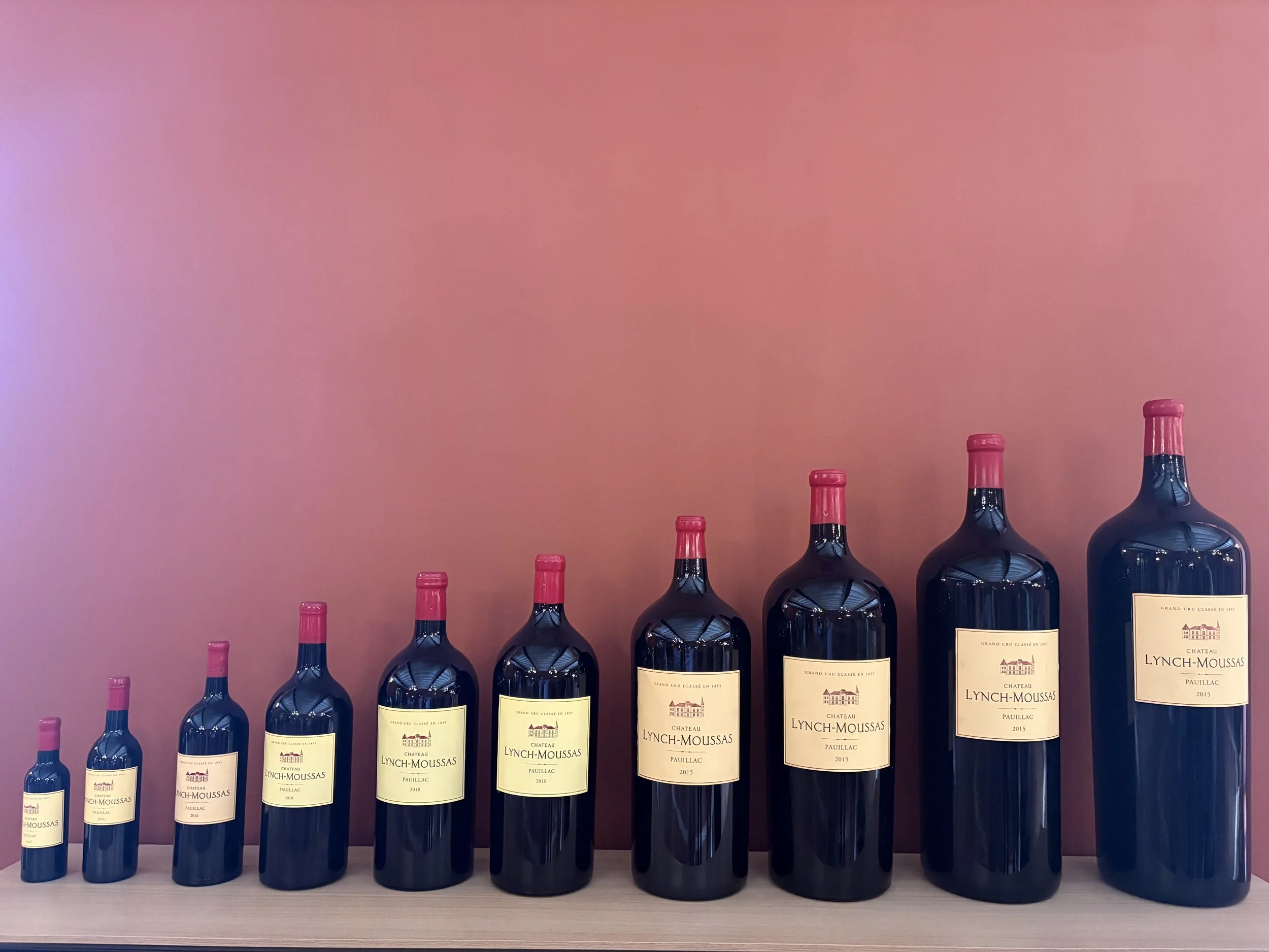 A collection of nine bottles of Château Lynch-Moussas wine, arranged from smallest to largest, on a white surface against a pink background.