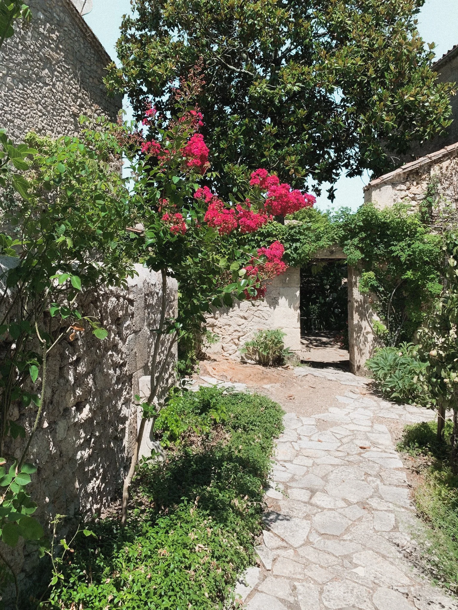 A stone pathway leading through a garden with pink flowering bushes and green foliage, with stone walls and an open wooden gate in the background.