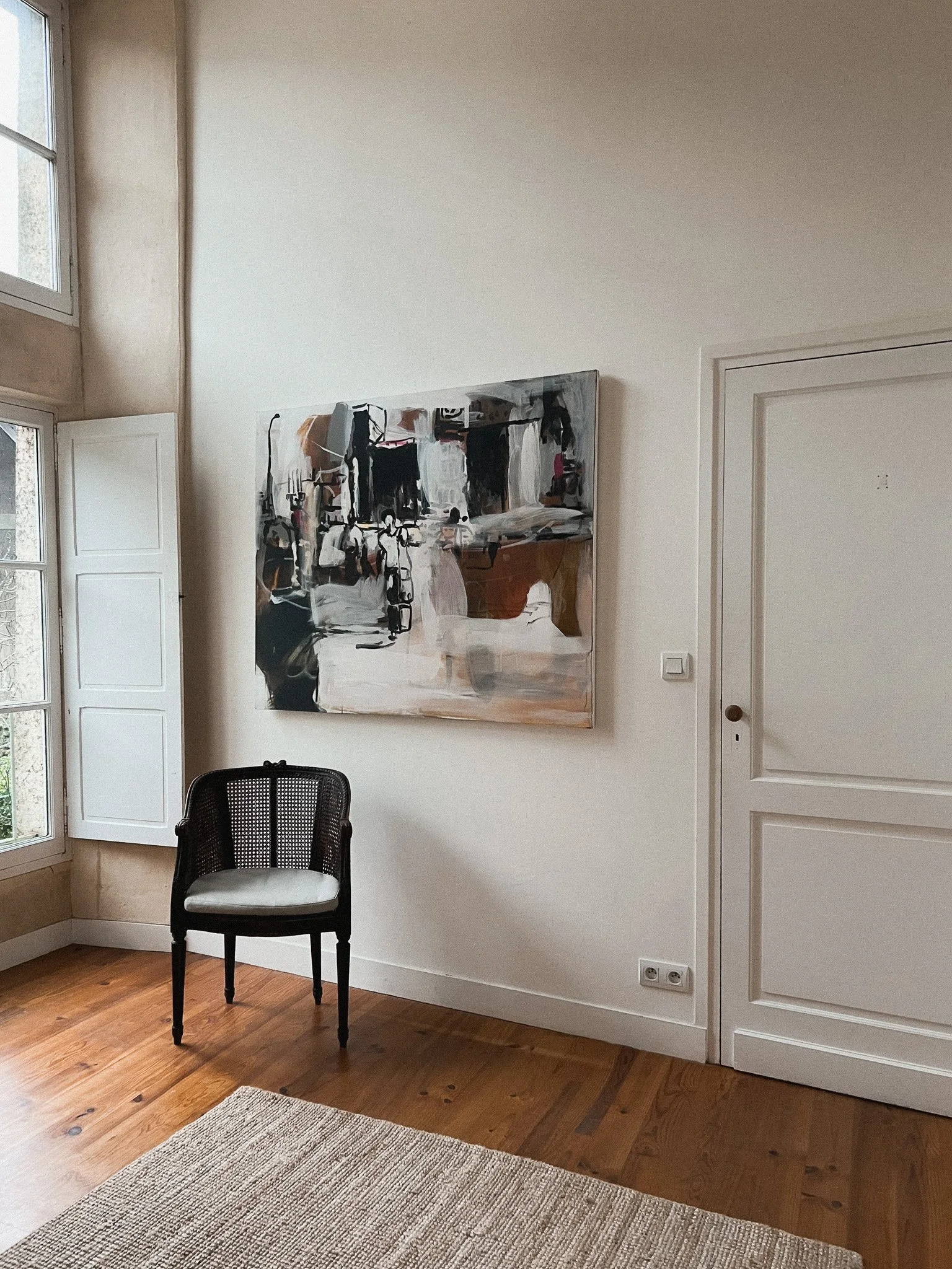 A room with white walls and wooden floors, featuring an abstract painting, a black chair with a white cushion, a window with white shutters, and a door.