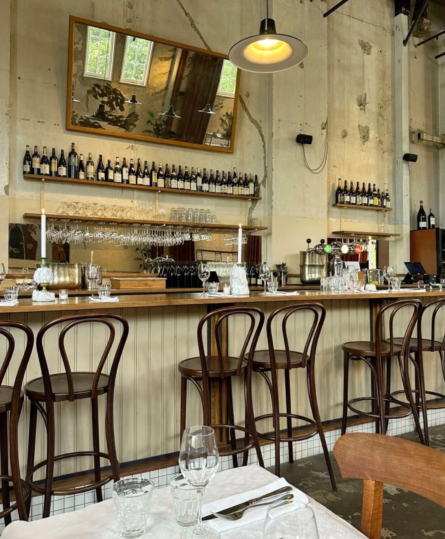 Interior of a restaurant bar with high wooden chairs, a table with glassware and utensils, and a rustic wall decorated with bottles and mirrors.