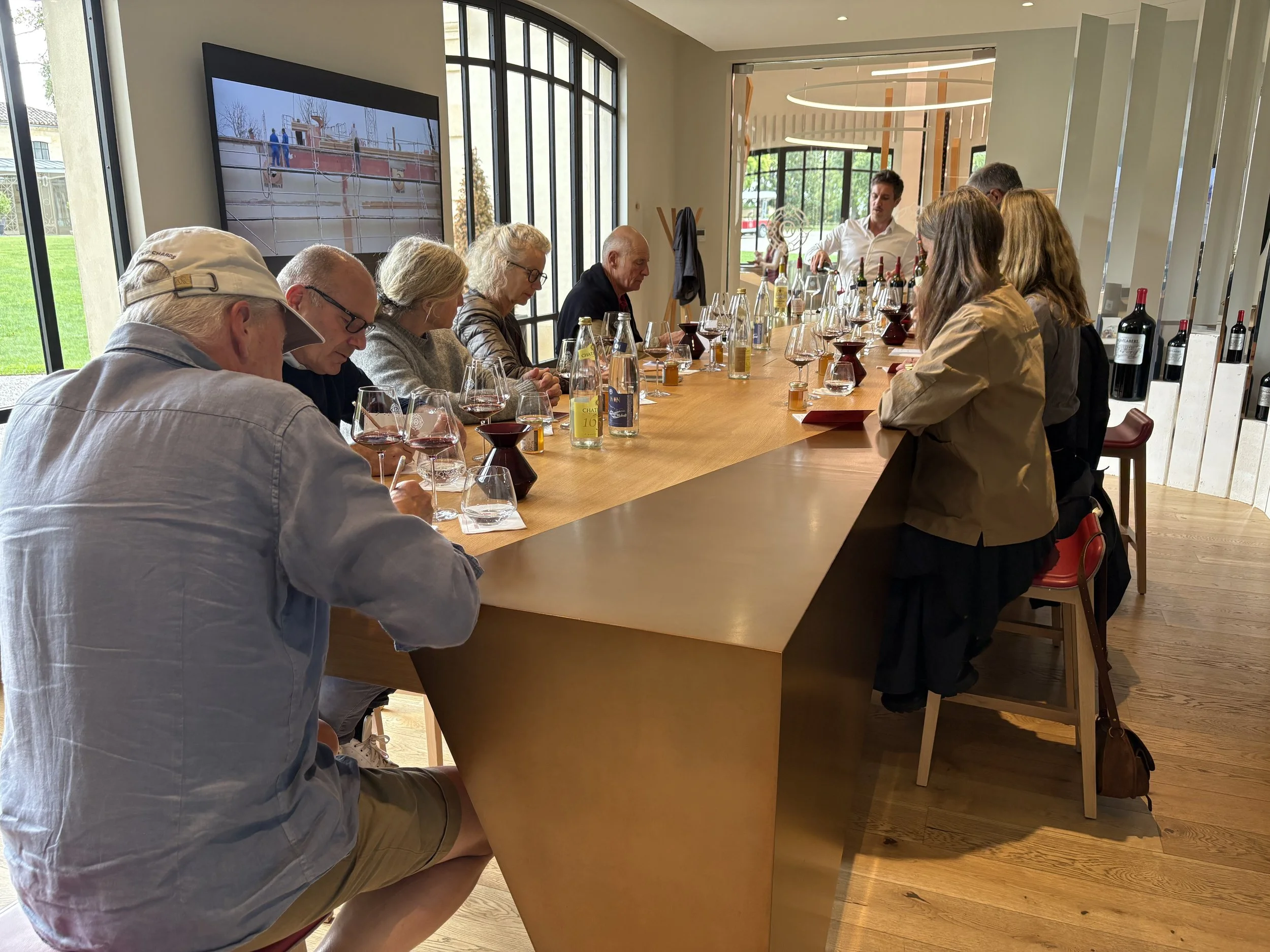 Group of people sitting at a long table in a modern, well-lit room, participating in a wine tasting event with wine glasses, bottles, and tasting notes in front of them.
