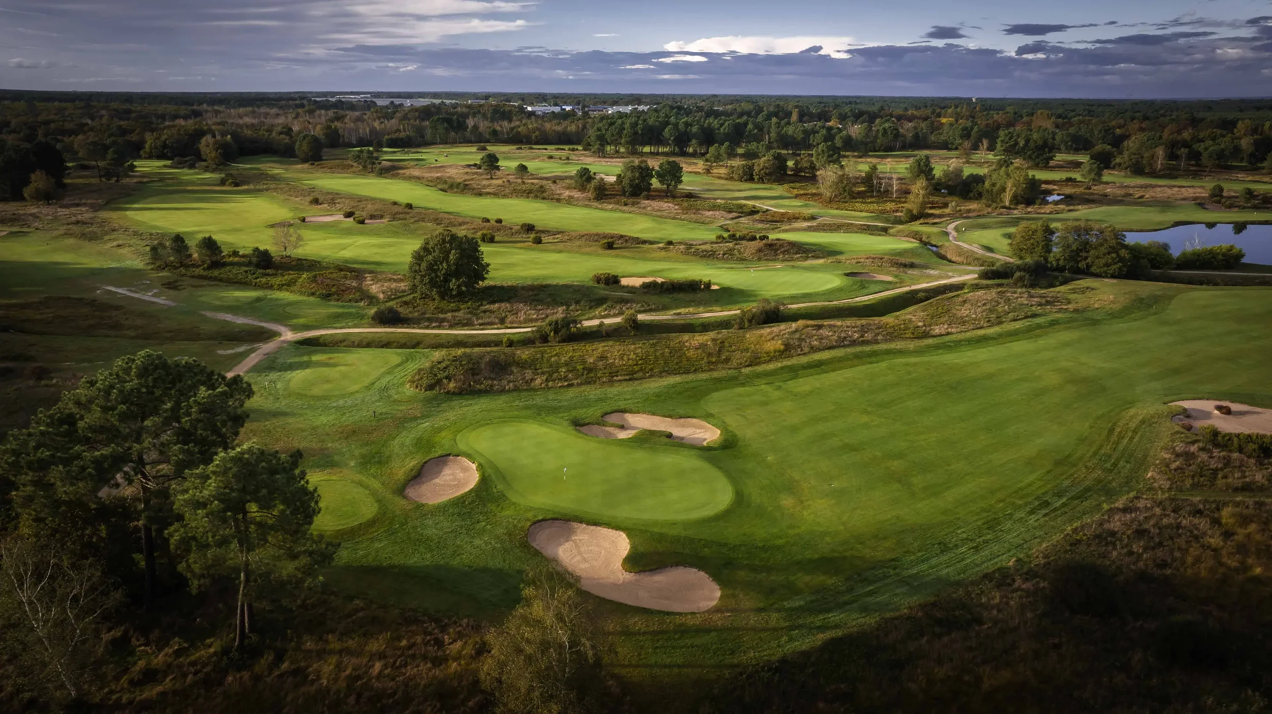 Aerial view of a golf course with green fairways, sand bunkers, and water hazards, surrounded by trees and open land under a cloudy sky.