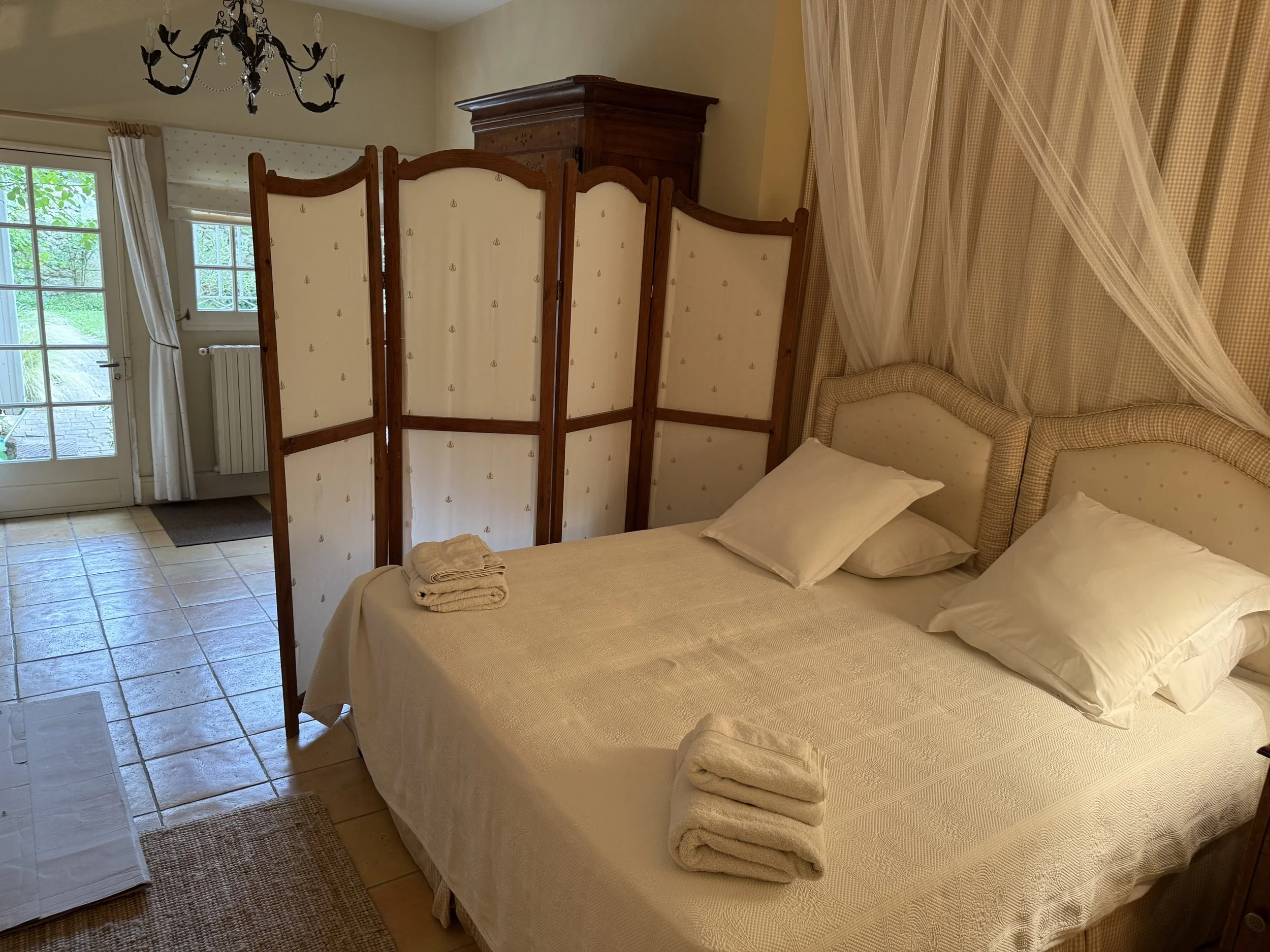 Bedroom with a large bed with pillows and folded towels, a privacy screen, a wooden wardrobe, a chandelier, a door with glass panels leading outside, and beige curtains.