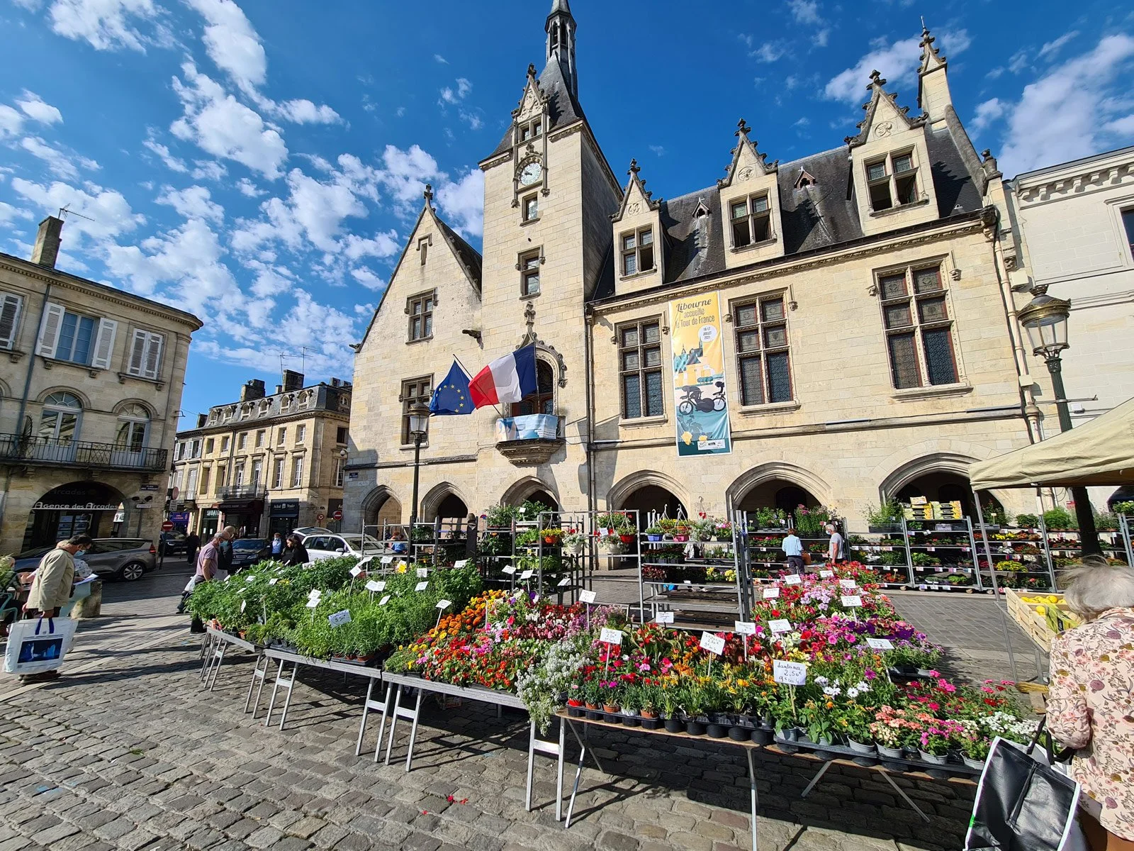 A historic stone building with a clock tower, French and European flags, and a colorful banner. In front, a flower market with various blooming plants and flowers on display, and people shopping on a cobblestone street under a partly cloudy blue sky.