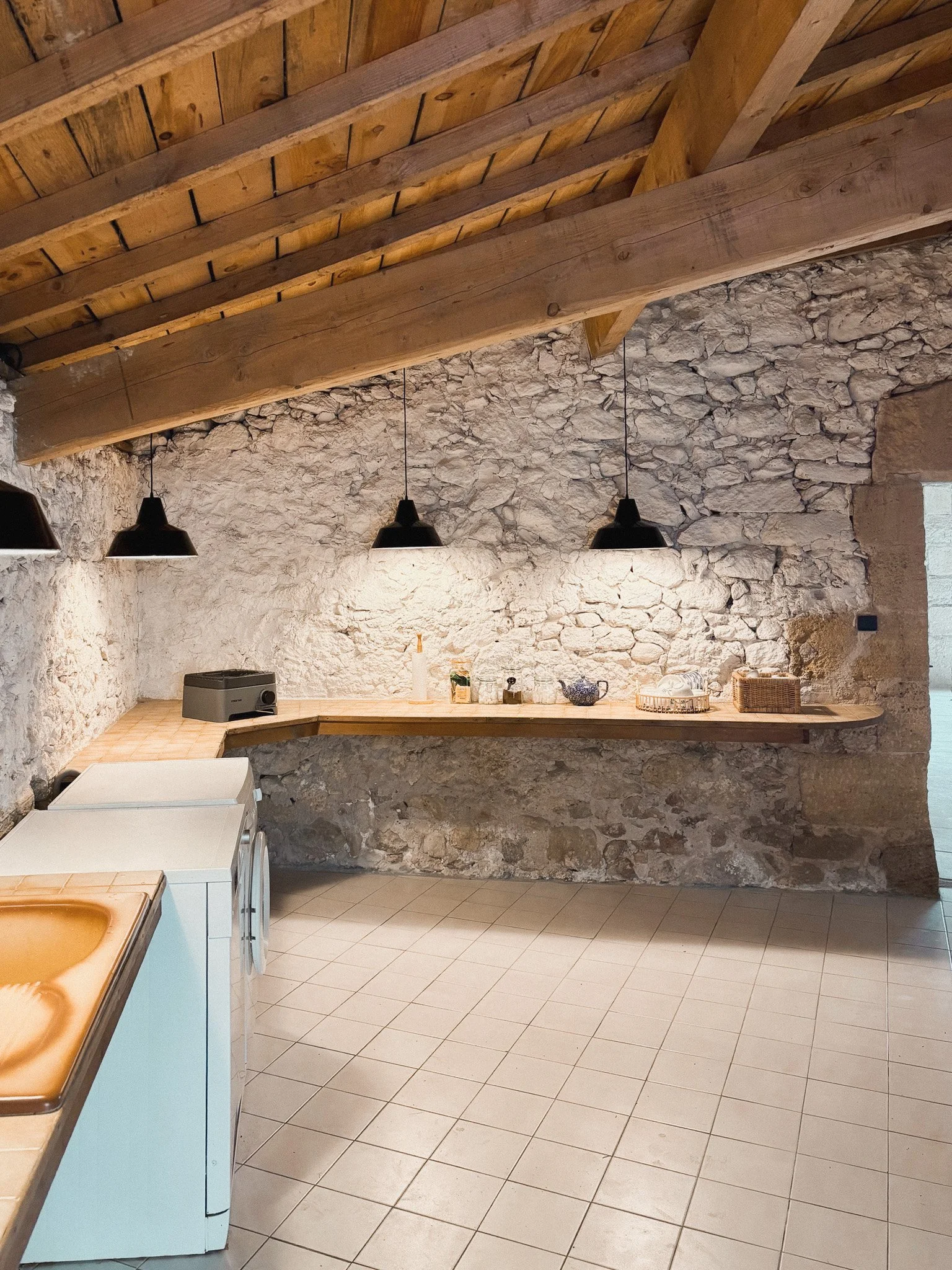 A rustic kitchen with stone walls, a wooden vaulted ceiling, black pendant lights, a wooden countertop, and a tiled floor. There is a washer, a small oven, and various kitchen items on the counter.