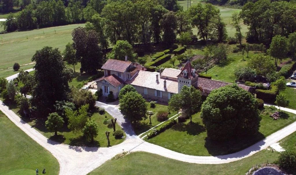 An aerial view of a large, historic house surrounded by lush green trees and landscaped gardens, with gravel driveways curving around the property.