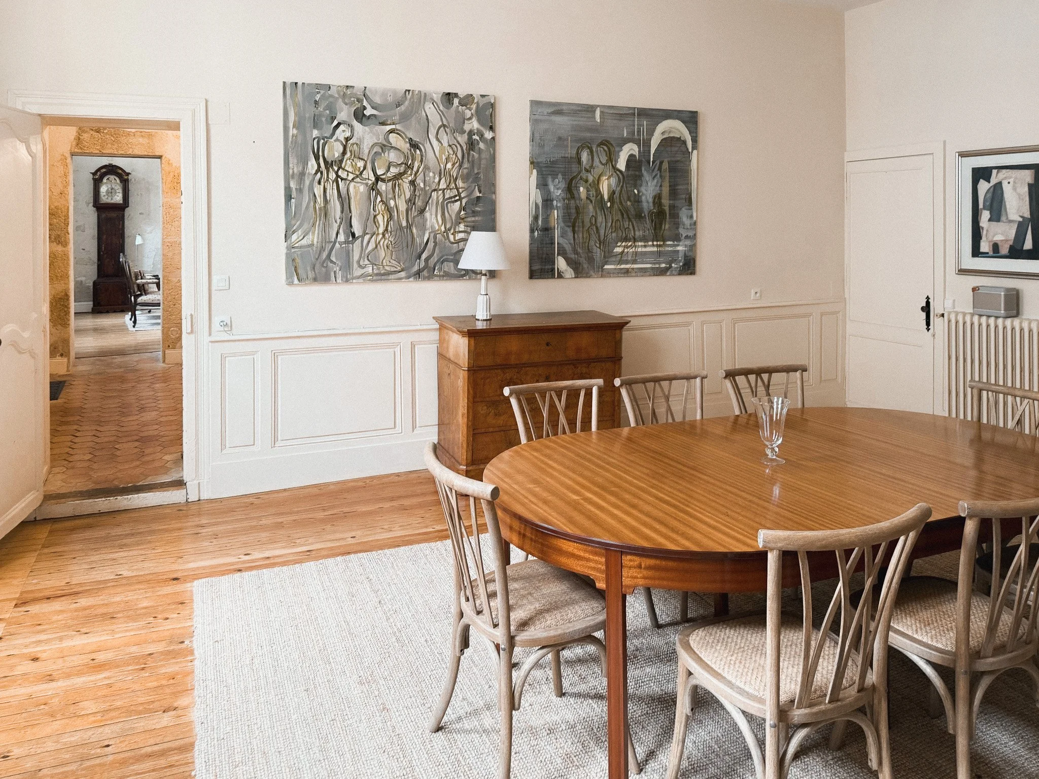 A dining room with a wooden table and six chairs, a wooden sideboard with a lamp, abstract artwork on the wall, a white wall with decorative paneling, a door, and a doorway leading to another room with an antique clock and chair.