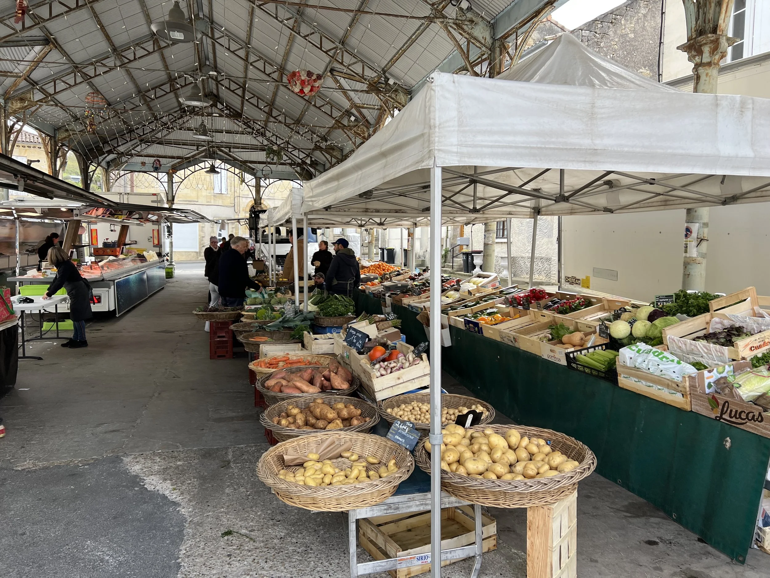 Open-air market with fresh vegetables and fruits under a metal and glass roof, customers shopping, and vendors attending to their stalls.