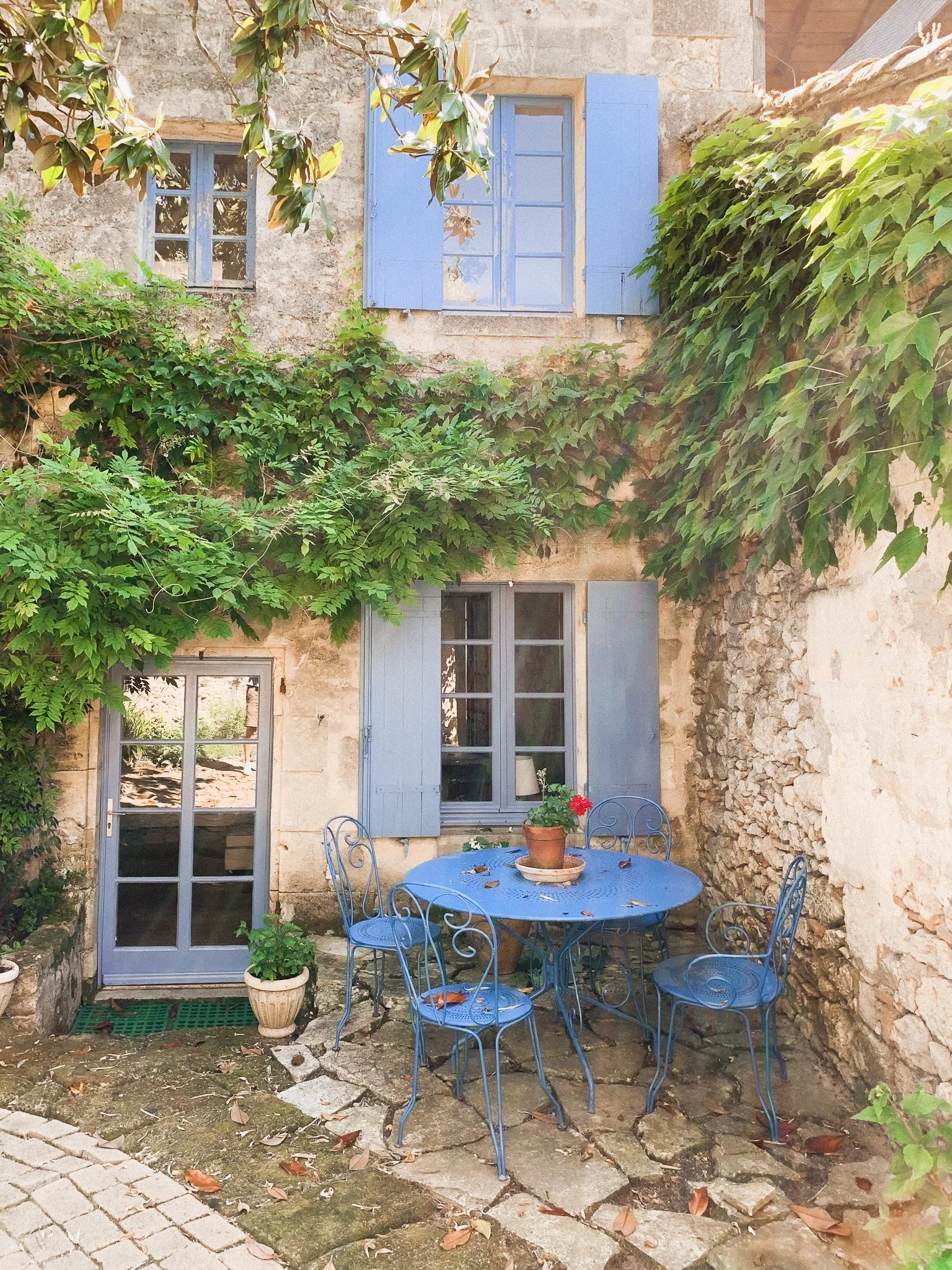 A cozy outdoor patio with a round blue metal table and four matching chairs, decorated with a potted red flower plant, set against a stone wall with green ivy and two windows with blue shutters.