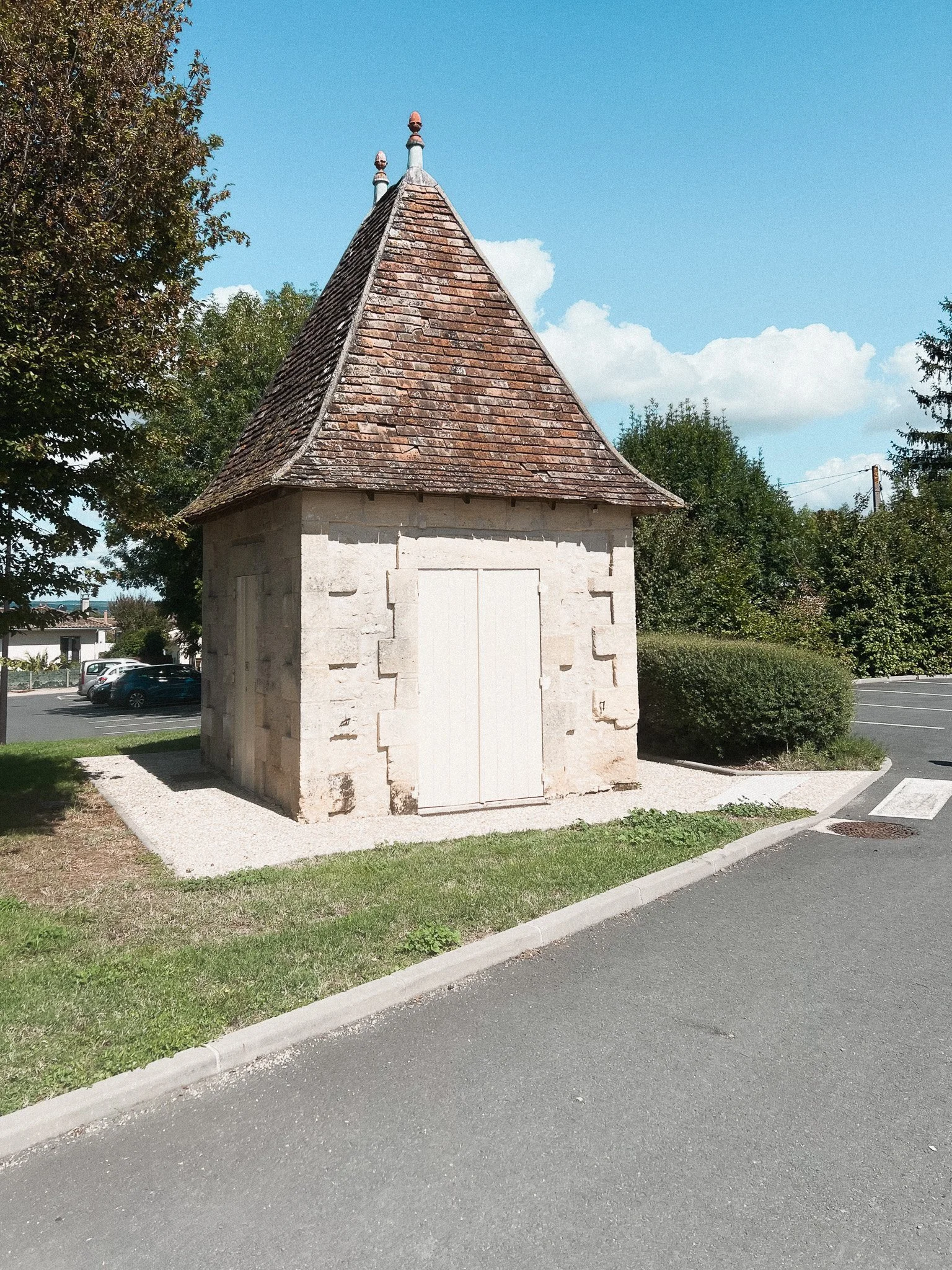 A small, stone building with a pointed, shingled roof in a parking lot, surrounded by greenery and a few parked cars.