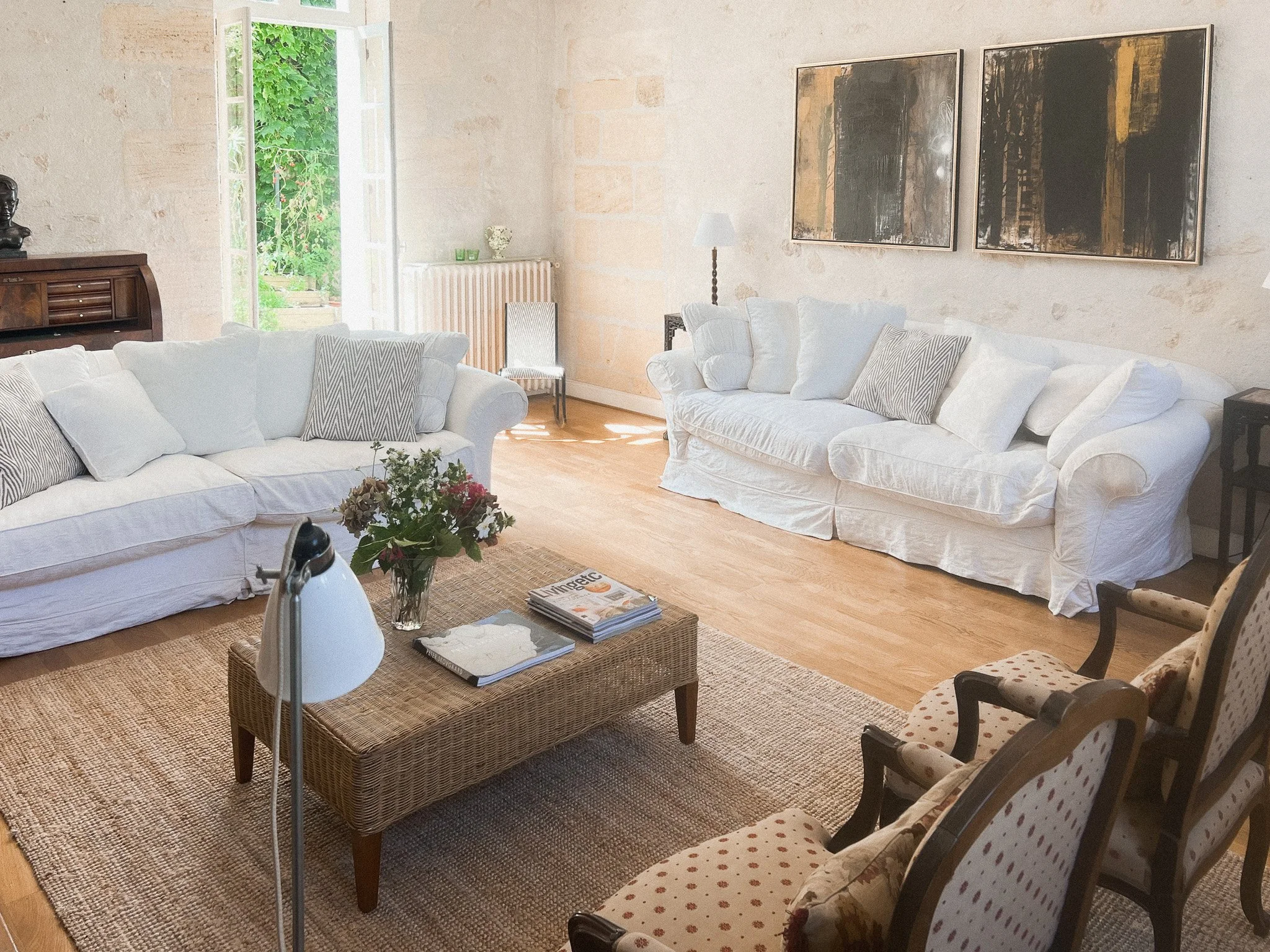 Bright living room with two white sofas, a wooden coffee table with magazines and a vase of flowers, a patterned armchair, and large artwork on the wall. Open door shows greenery outside.