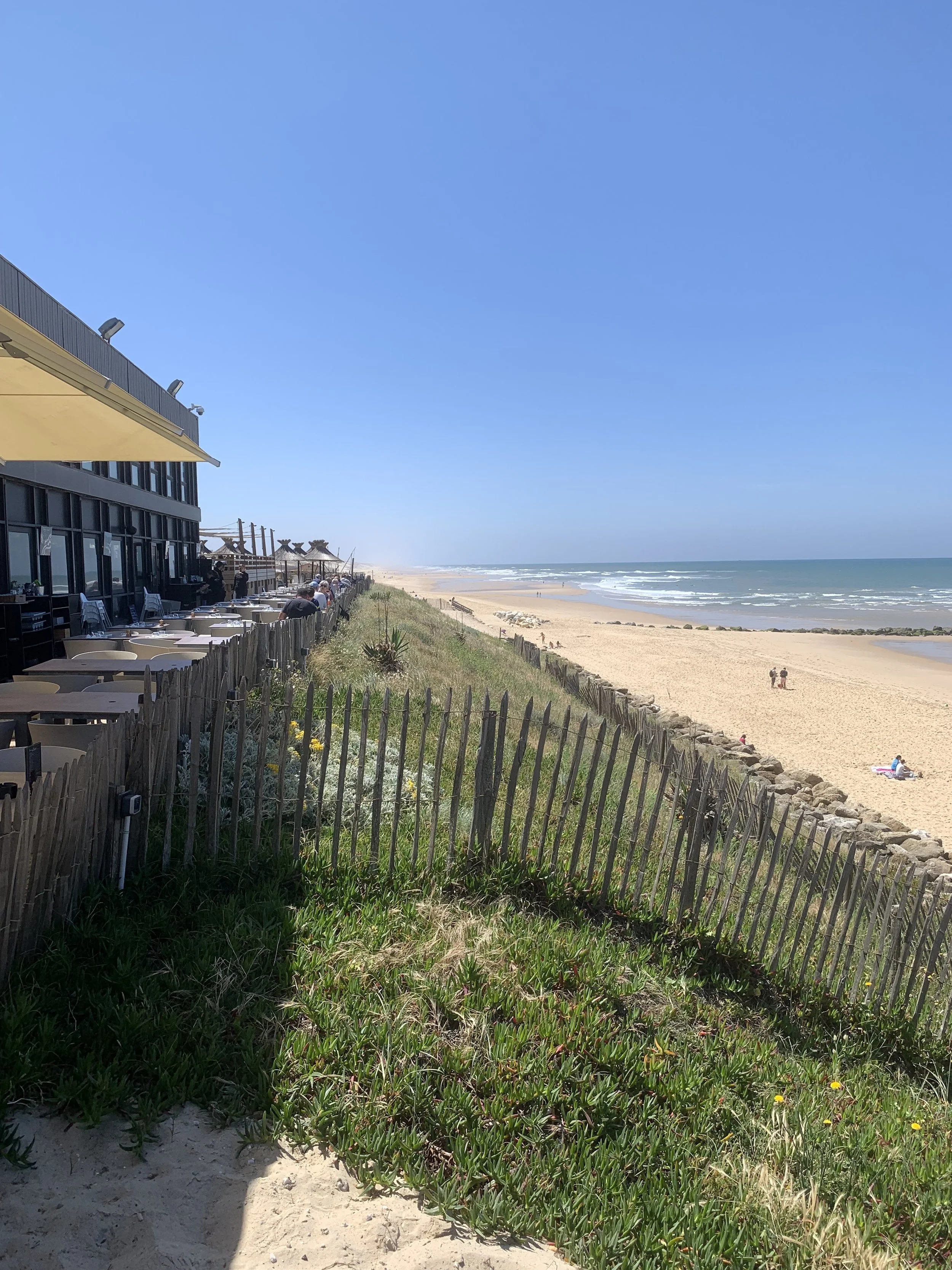 View of a sandy beach with people, blue sky, ocean waves, a beachfront restaurant with outdoor tables, and a wooden fence separating the restaurant area from the beach.