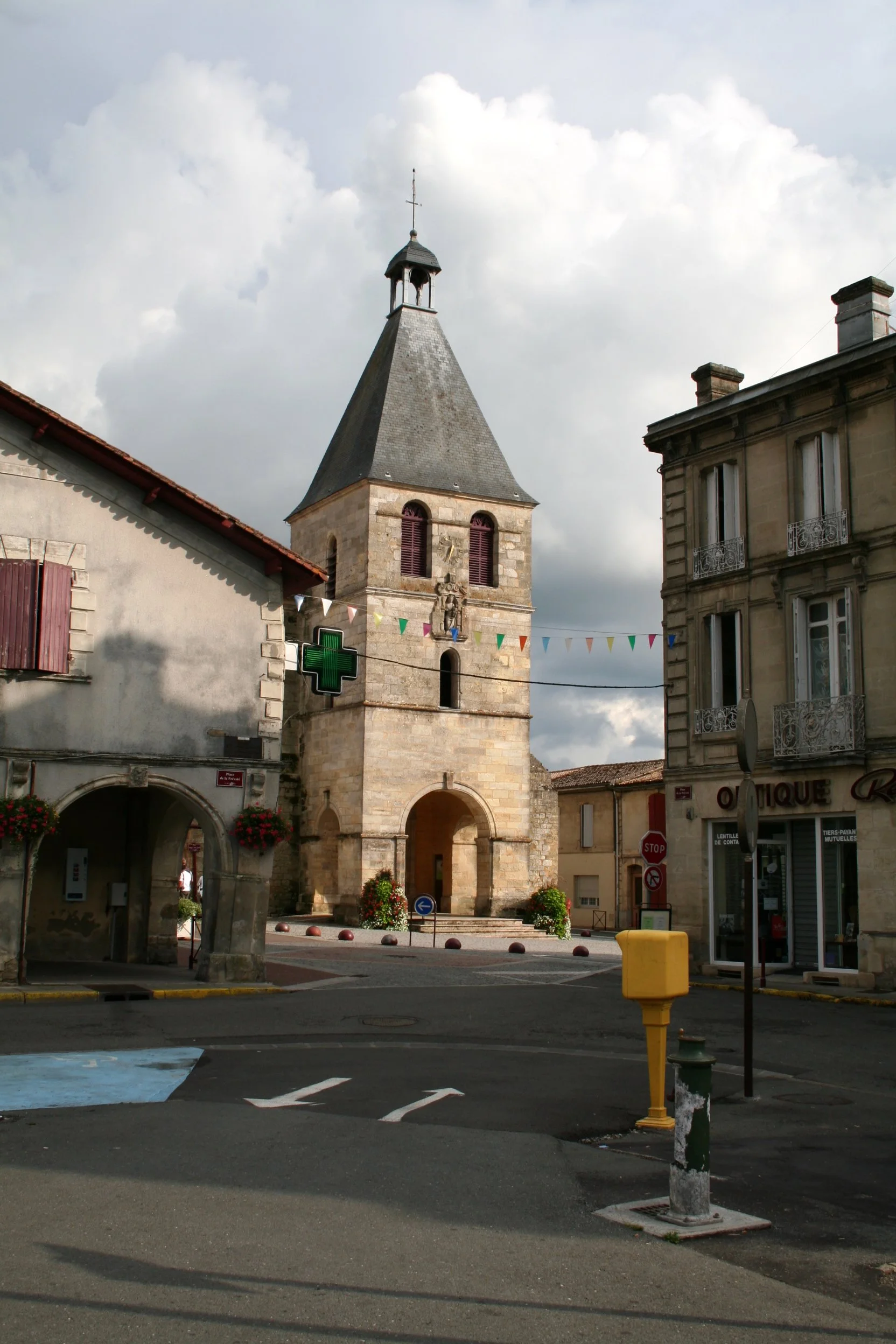 A scenic view of a small European town square with a historic stone clock tower or church with a steep roof, surrounded by older buildings, colorful bunting, and a pharmacy sign, under a partly cloudy sky.