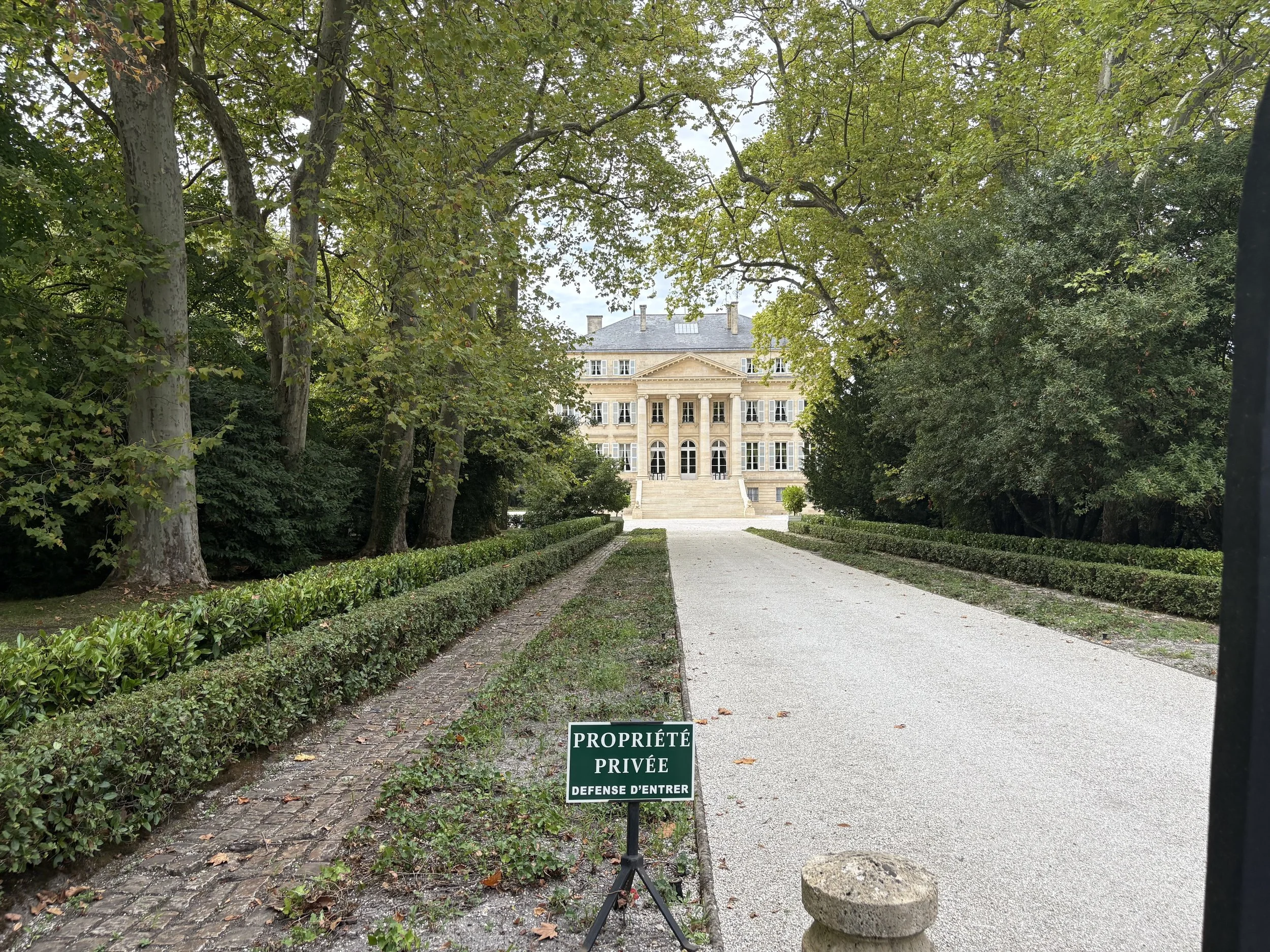 A view of a large, elegant house through trees, with a gravel pathway leading to the house and a sign in French and English that reads 'Private Property. No Entry.'